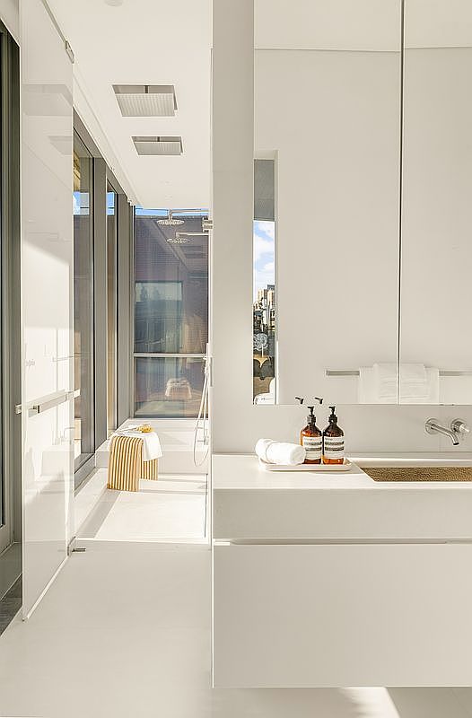 This minimalist, modern bathroom features a sleek, wall-mounted vanity with a stone countertop and a recessed sink. A large mirror reflects the bright, airy space, which leads into a shower area with floor-to-ceiling glass panels and a view of the city skyline. The clean lines and neutral color palette create a sophisticated, spa-like atmosphere.