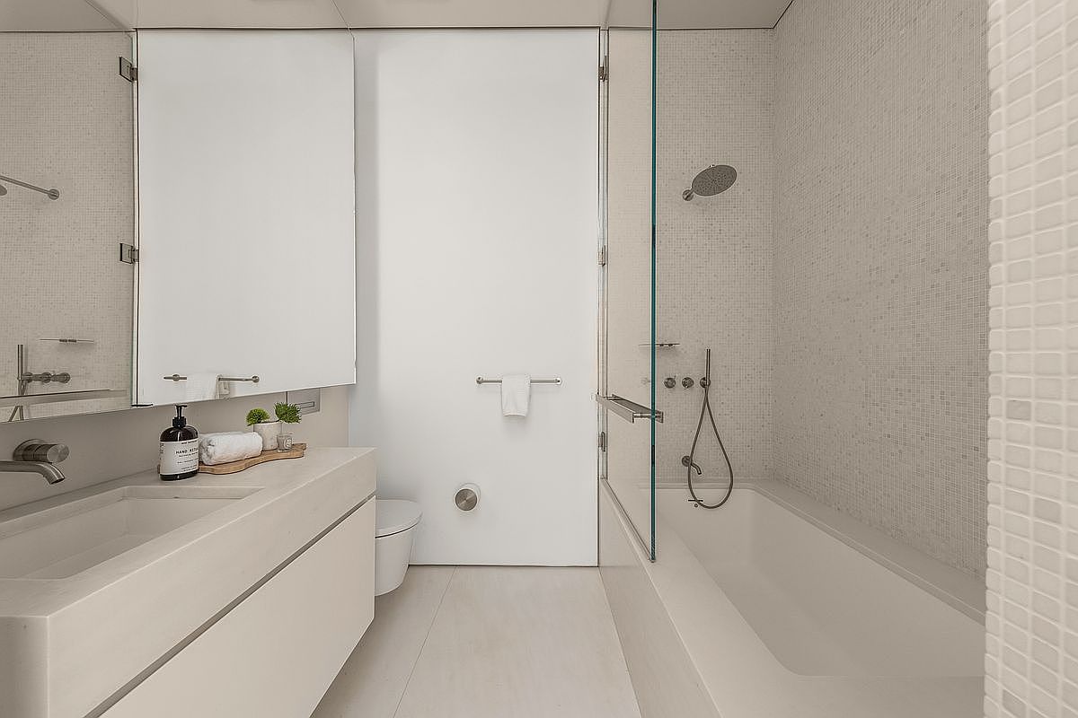 This modern, minimalist bathroom features a sleek vanity with a stone countertop and a wall-mounted faucet. To the right, a glass-enclosed shower and tub combination is finished with small, light-colored square tiles. The space is bright and clean, utilizing a neutral palette and contemporary fixtures to create a spa-like atmosphere.