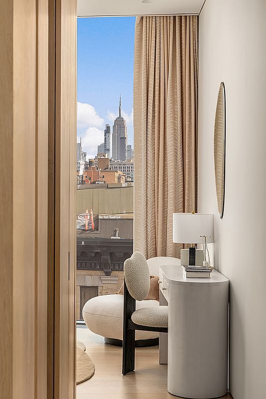 This intimate bedroom space features a sleek, curved white desk paired with a textured, rounded chair, creating a sophisticated nook. A tall window with floor-to-ceiling drapery frames a striking view of the city skyline, including the Empire State Building. The room is accented by a minimalist wall mirror and warm, neutral tones, offering a serene and elevated urban atmosphere.