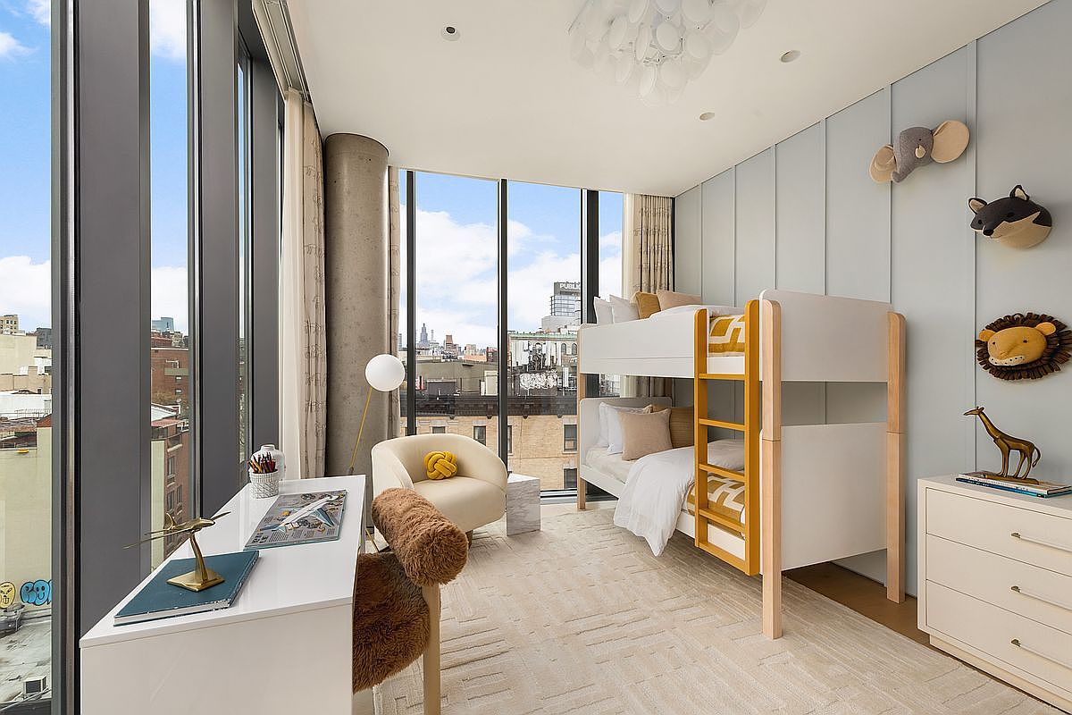 This bright and modern children's bedroom features a light wood bunk bed set against a soft blue paneled wall adorned with whimsical animal-themed wall decor. A sleek white desk with a textured chair sits near expansive floor-to-ceiling windows that provide an urban cityscape view, while a cozy armchair and a light-toned dresser complete the functional layout. The room is illuminated by a unique, cloud-like pendant light, creating a playful yet sophisticated atmosphere.