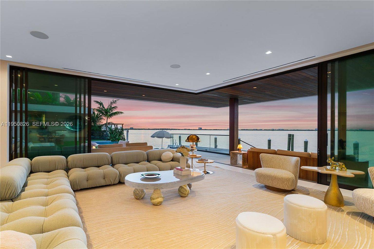This is a luxurious living room featuring a large, modular sofa, a unique coffee table, and plush seating. The room opens to an outdoor area with water views, creating a seamless indoor-outdoor living experience. The design is modern and sophisticated, emphasizing comfort and elegance.