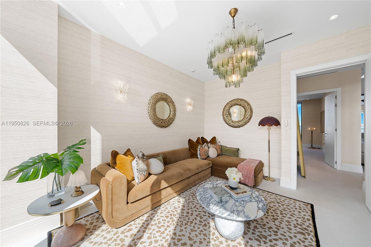 This interior shot showcases a stylish living room featuring a plush brown sectional sofa adorned with decorative pillows, complemented by a unique marble-topped coffee table and a patterned rug. The room is illuminated by a statement chandelier and wall sconces, enhancing the textured wallpaper and creating a warm, inviting atmosphere. The open doorway leads to another room, suggesting a spacious and well-designed home layout.