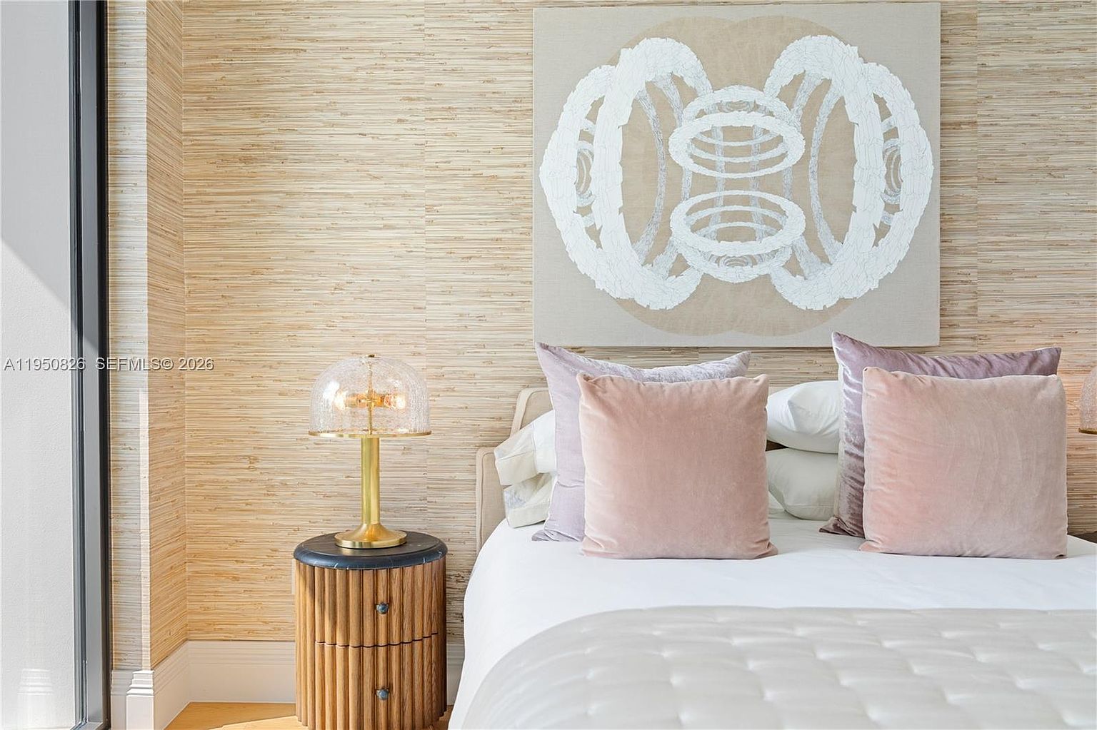 This is a bedroom featuring a bed with pink and lavender pillows, a nightstand with a lamp, and a piece of art hanging above the bed. The walls are covered in a textured wallpaper, adding depth and warmth to the room. The overall impression is one of comfort and style, creating a relaxing and inviting space.