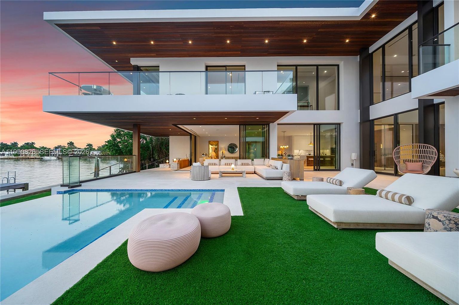 This image showcases the rear exterior of a luxurious modern home, emphasizing the outdoor living space. A sleek pool is the focal point, complemented by stylish lounge chairs and plush seating on an artificial lawn. The architecture features clean lines, expansive glass doors, and a cantilevered balcony, creating a seamless transition between indoor and outdoor living.