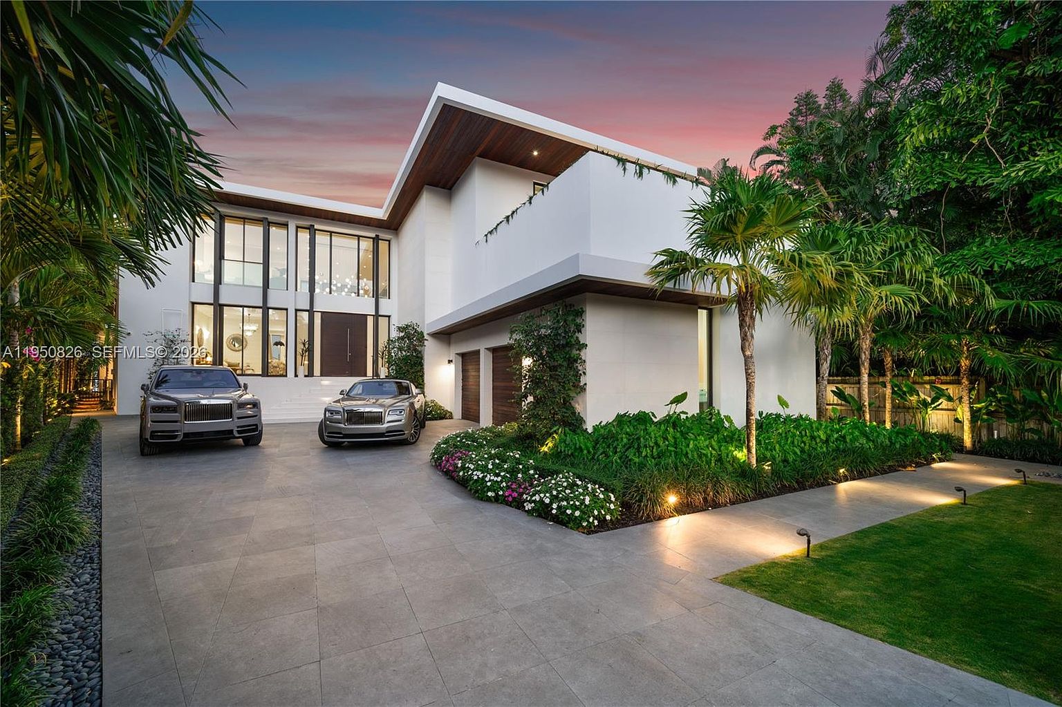This is a front exterior view of a modern, luxurious home. The house features a striking architectural design with clean lines, a flat roof, and a combination of white and wood accents. Two high-end cars are parked in the driveway, and the landscaping includes lush greenery, flowers, and palm trees, enhancing the property's curb appeal.