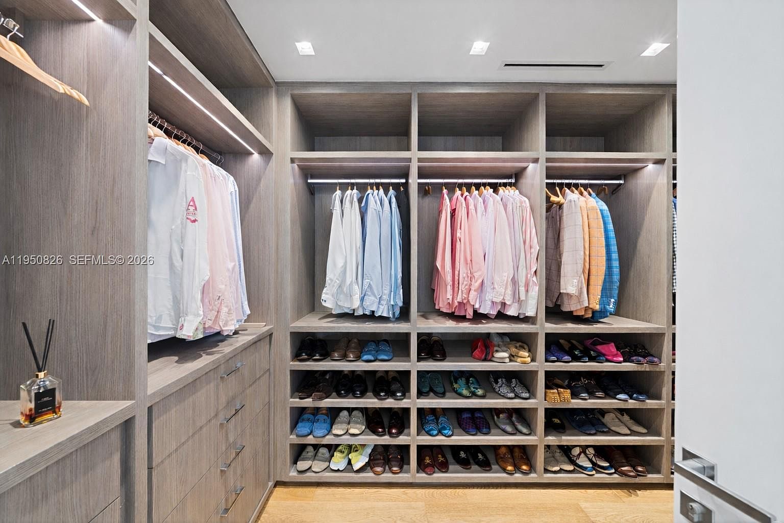 This is a well-organized walk-in closet featuring custom wood shelving and drawers. The closet is filled with neatly hung shirts and rows of shoes, showcasing ample storage space. The lighting is bright and modern, enhancing the overall luxurious feel of the closet.
