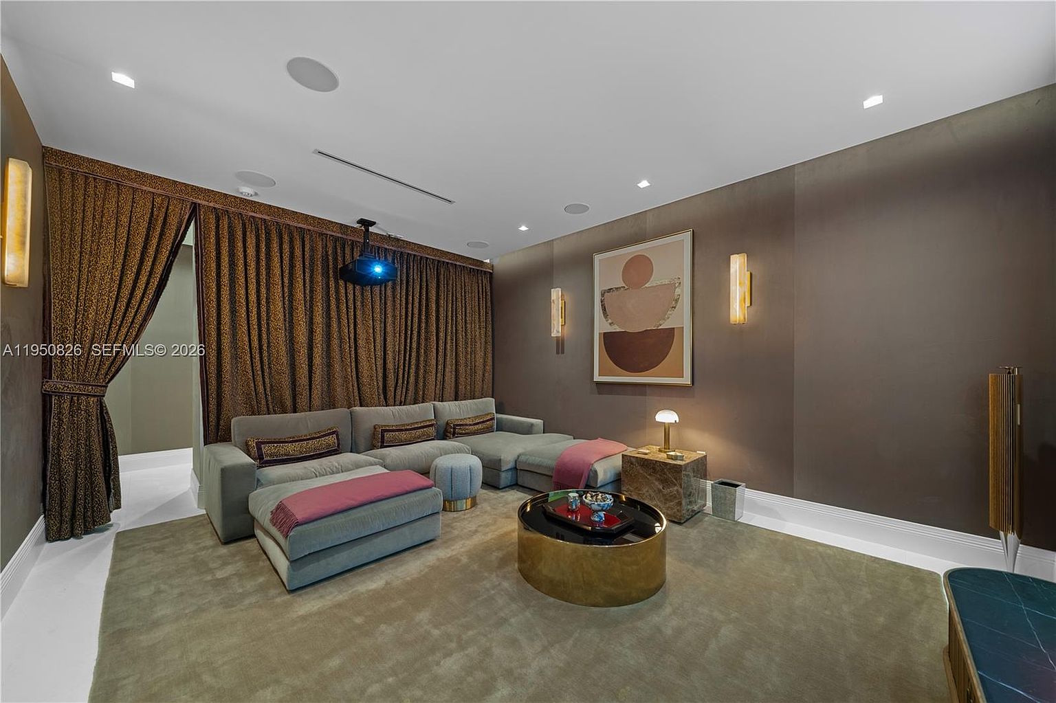 This is an interior shot of a living room or home theater, featuring a large sectional sofa with throw pillows and blankets, a round coffee table, and a projector mounted on the ceiling. The room is decorated with brown walls, curtains, and artwork, creating a cozy and cinematic atmosphere. The lighting is soft and warm, enhancing the room's inviting ambiance.
