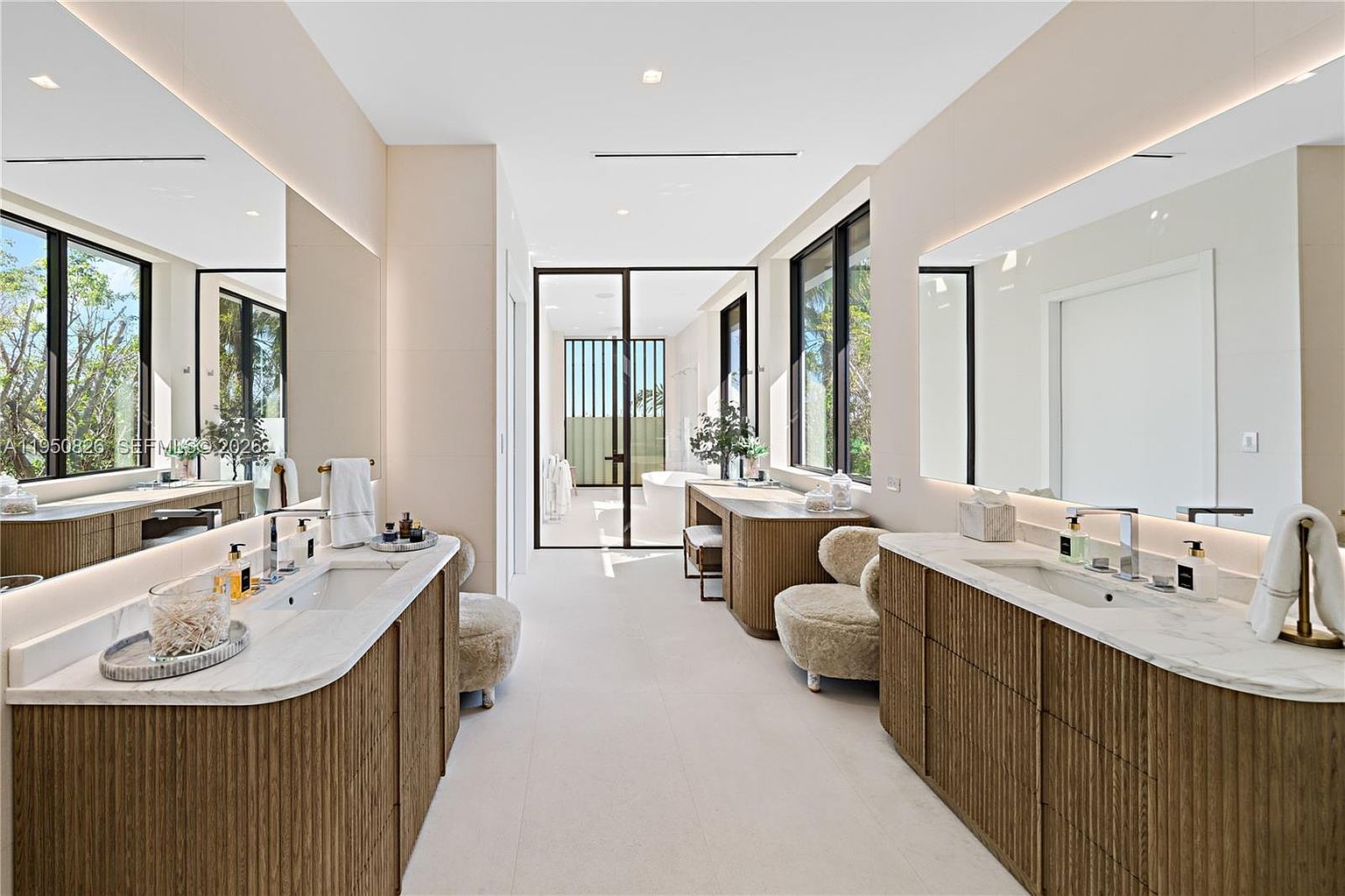 This is a luxurious primary bathroom featuring dual vanities with ribbed wood cabinetry and marble countertops. Large mirrors and windows provide ample light, enhancing the spacious feel. A separate bathing area is visible through a glass door, contributing to the spa-like atmosphere.