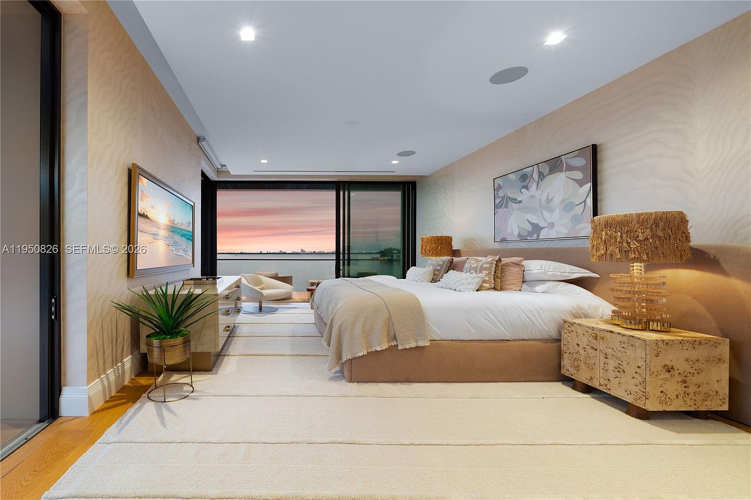 This is a luxurious primary bedroom featuring a large bed with multiple pillows and a throw blanket. The room has a neutral color palette with textured walls and a large piece of artwork above the bed. A sliding glass door leads to a balcony with a view of the water and a beautiful sunset, enhancing the room's serene and upscale ambiance.
