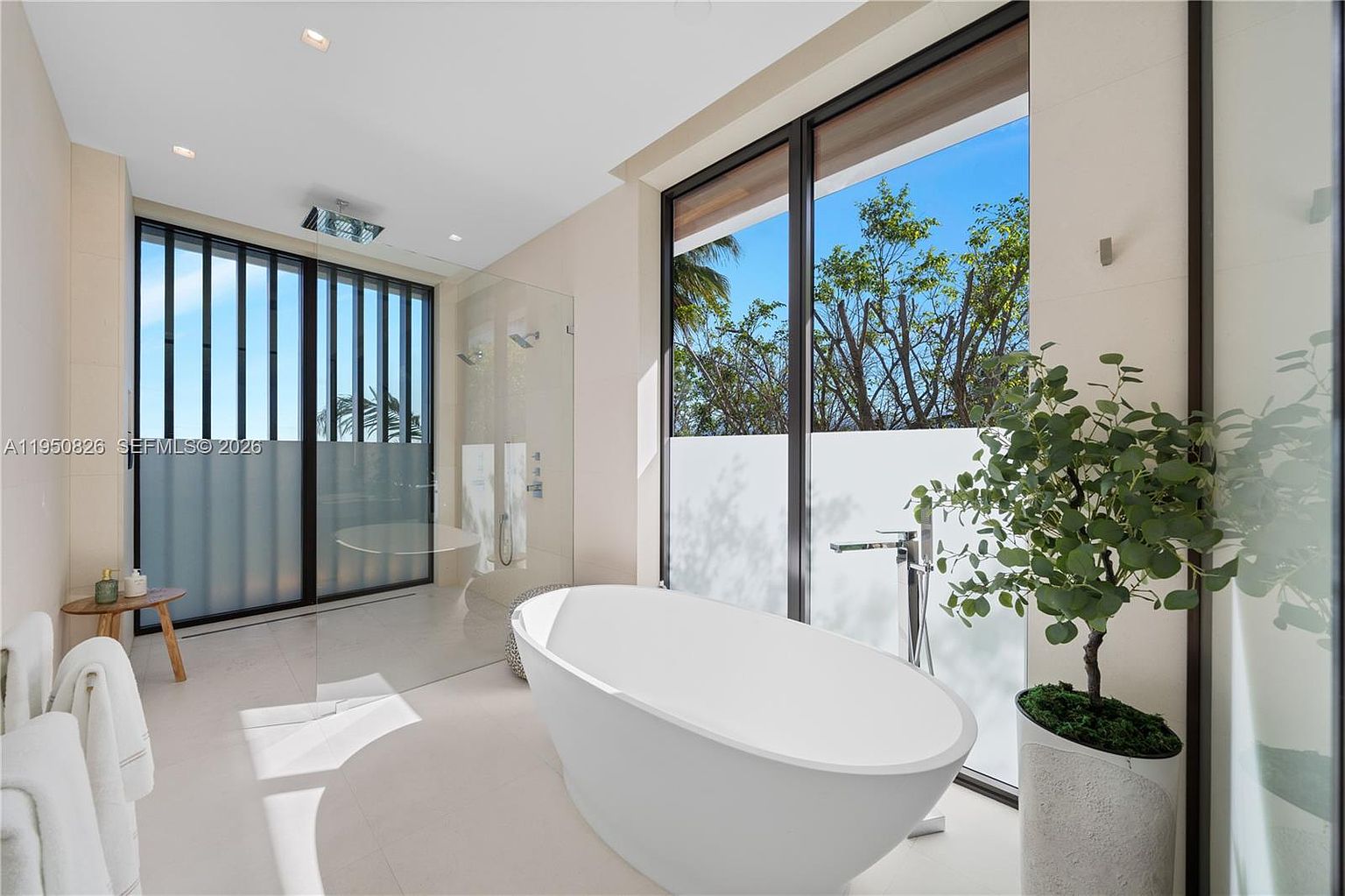 This is a luxurious primary bathroom featuring a freestanding white bathtub positioned near a large window offering natural light and a view of greenery. The space includes a glass-enclosed shower with dual showerheads and sleek fixtures. The overall design is modern and minimalist, emphasizing clean lines and a spa-like atmosphere.