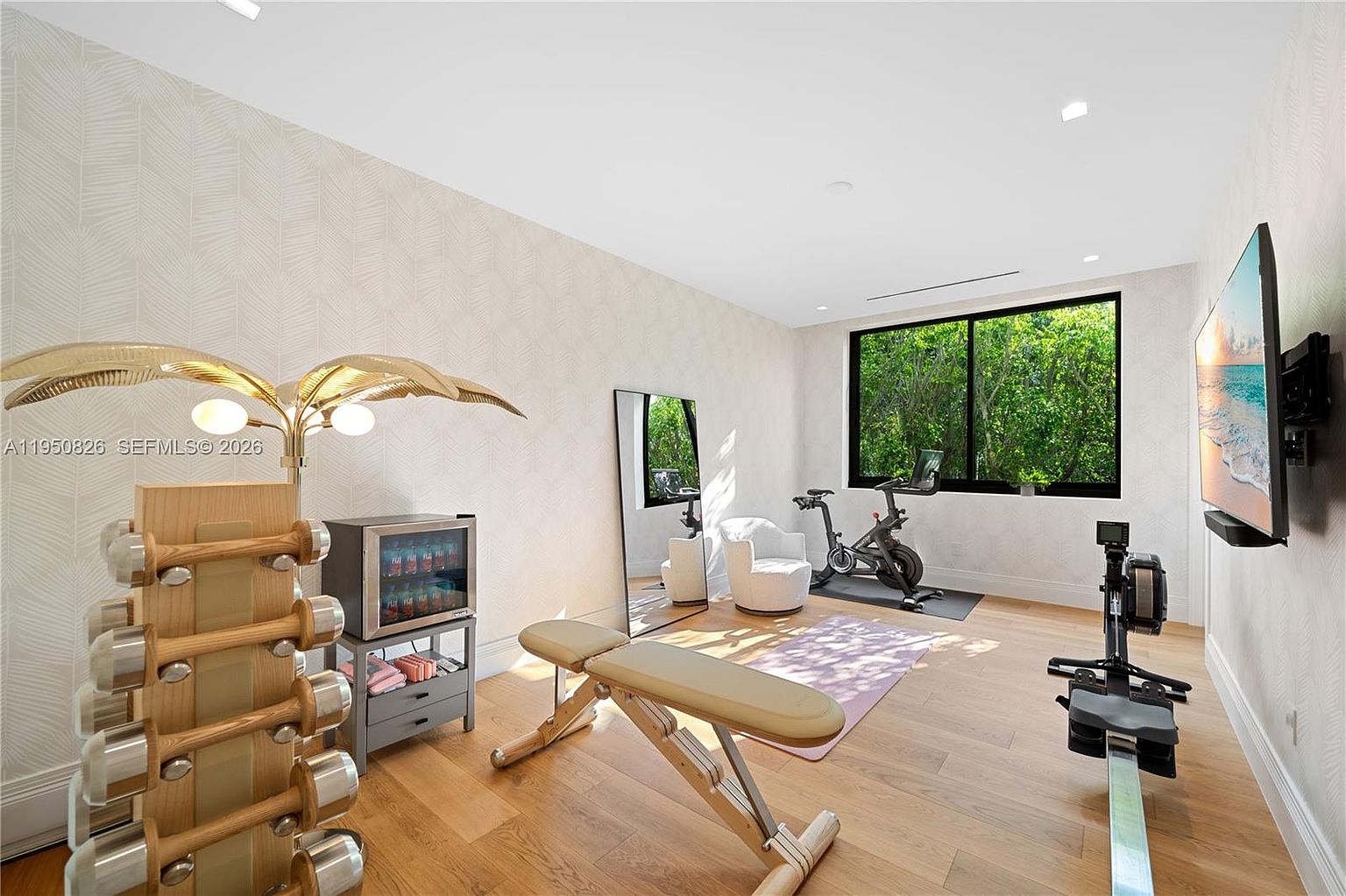 This is a well-equipped home gym featuring hardwood floors and stylish wallpaper. The room includes a weight rack, mini-fridge, adjustable bench, exercise bike, rowing machine, and a wall-mounted television. A large window provides natural light and a view of lush greenery, creating an inviting workout space.