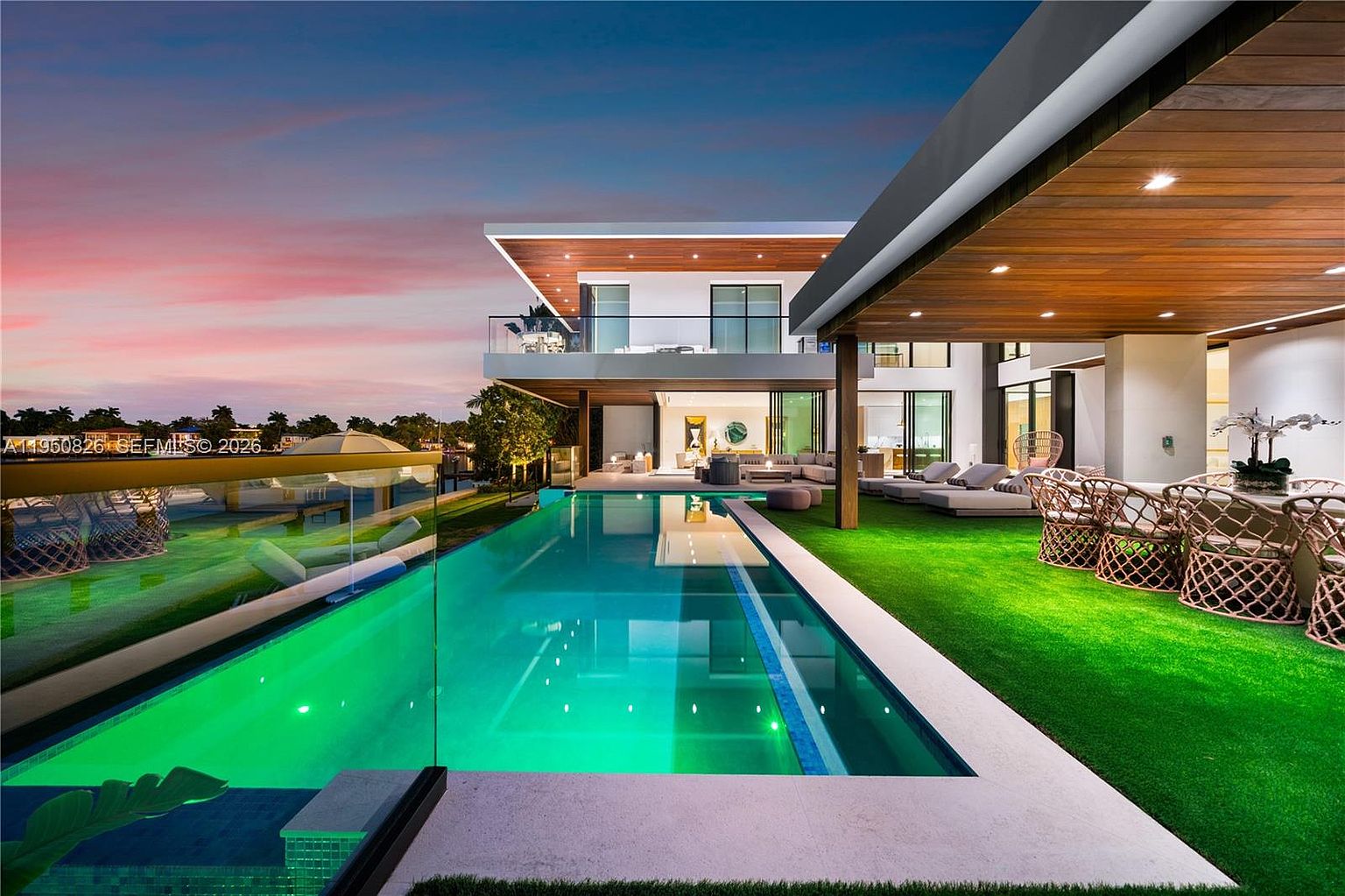 This image showcases a luxurious backyard featuring a modern two-story house with a sleek design. A long, rectangular pool with illuminated green water is the focal point, complemented by a well-manicured artificial lawn and outdoor seating areas. The scene is set during twilight, with a colorful sky adding to the overall ambiance of upscale living and relaxation.