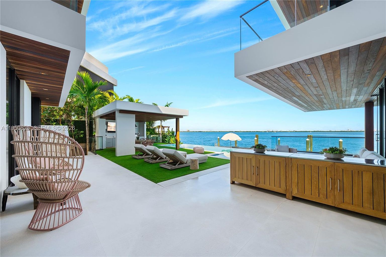 This image showcases a luxurious outdoor patio area with a stunning waterfront view. The space features modern architectural elements, including a covered lounge area, a built-in outdoor kitchen, and stylish lounge chairs arranged on artificial turf. The overall impression is one of high-end relaxation and sophisticated outdoor living.