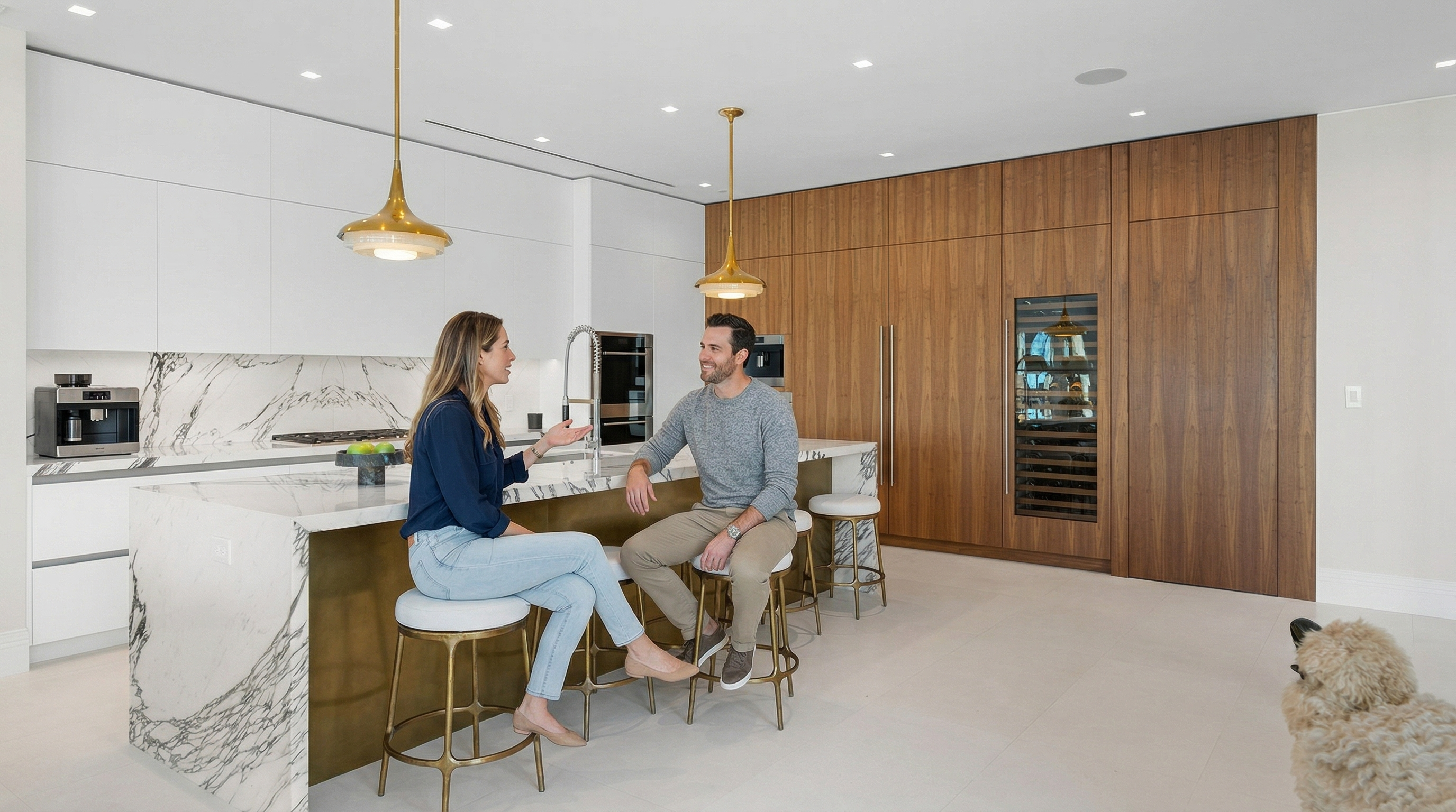 This is a bright and modern kitchen featuring a large island with a marble countertop and gold accents, complemented by white cabinetry and a wood-paneled wall with a built-in wine fridge. The kitchen is illuminated by pendant lights, and the overall design is sleek and luxurious. The perspective is from a low angle, emphasizing the size and features of the kitchen.