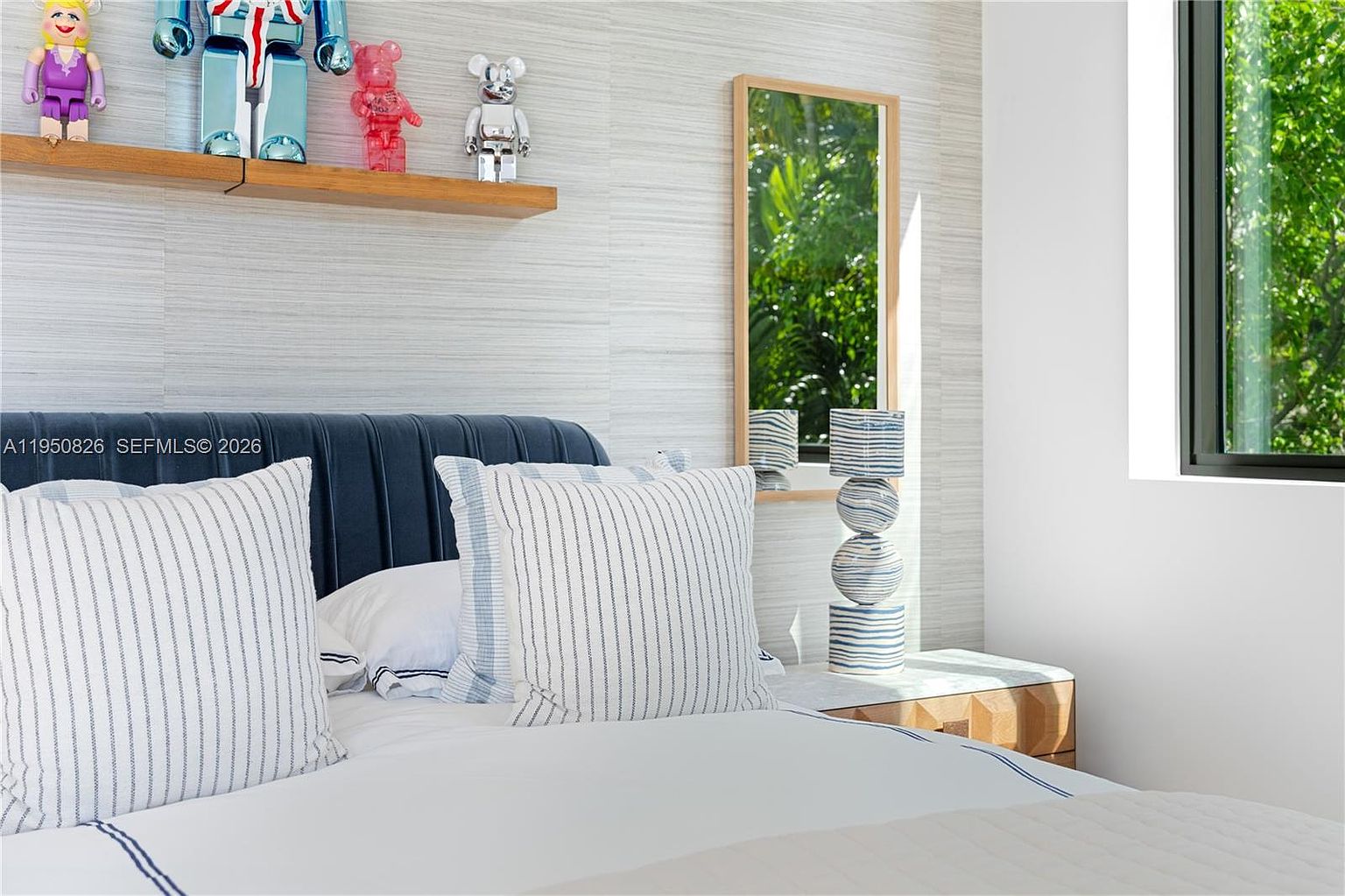 This is a bedroom featuring a bed with striped pillows and a blue headboard. A wooden shelf displays decorative figures, and a mirror reflects greenery. The room has a modern and clean aesthetic, with a neutral color palette and natural light coming through the window.