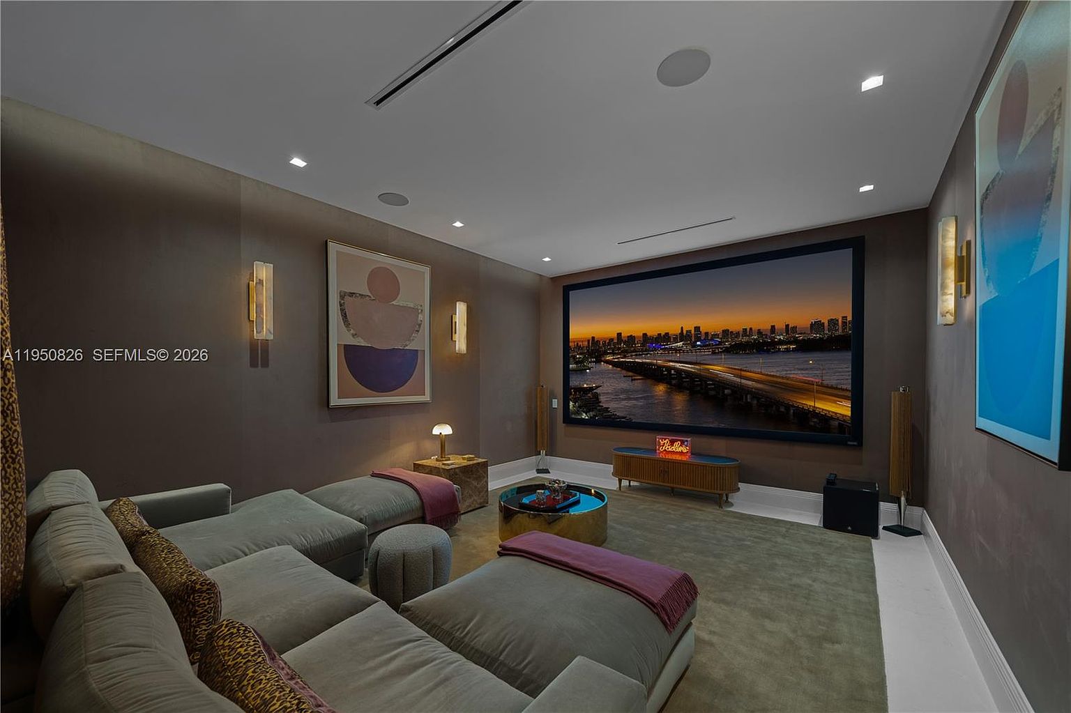 This is a luxurious home theater room featuring a large screen displaying a cityscape at dusk, comfortable sectional seating with decorative pillows, and ambient lighting from wall sconces. The room is decorated with modern art and has a sophisticated color palette, creating a cozy and high-end entertainment space. The perspective is from the back of the room, looking towards the screen.