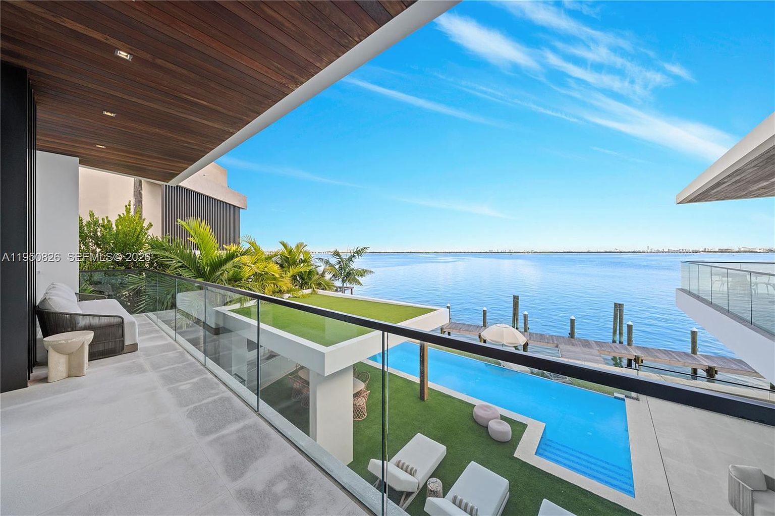 This image showcases a luxurious outdoor patio/deck/balcony area with stunning waterfront views. The space features modern design elements, including a glass railing, sleek furniture, and a swimming pool with an adjacent lounge area. The overall impression is one of high-end relaxation and sophisticated outdoor living.