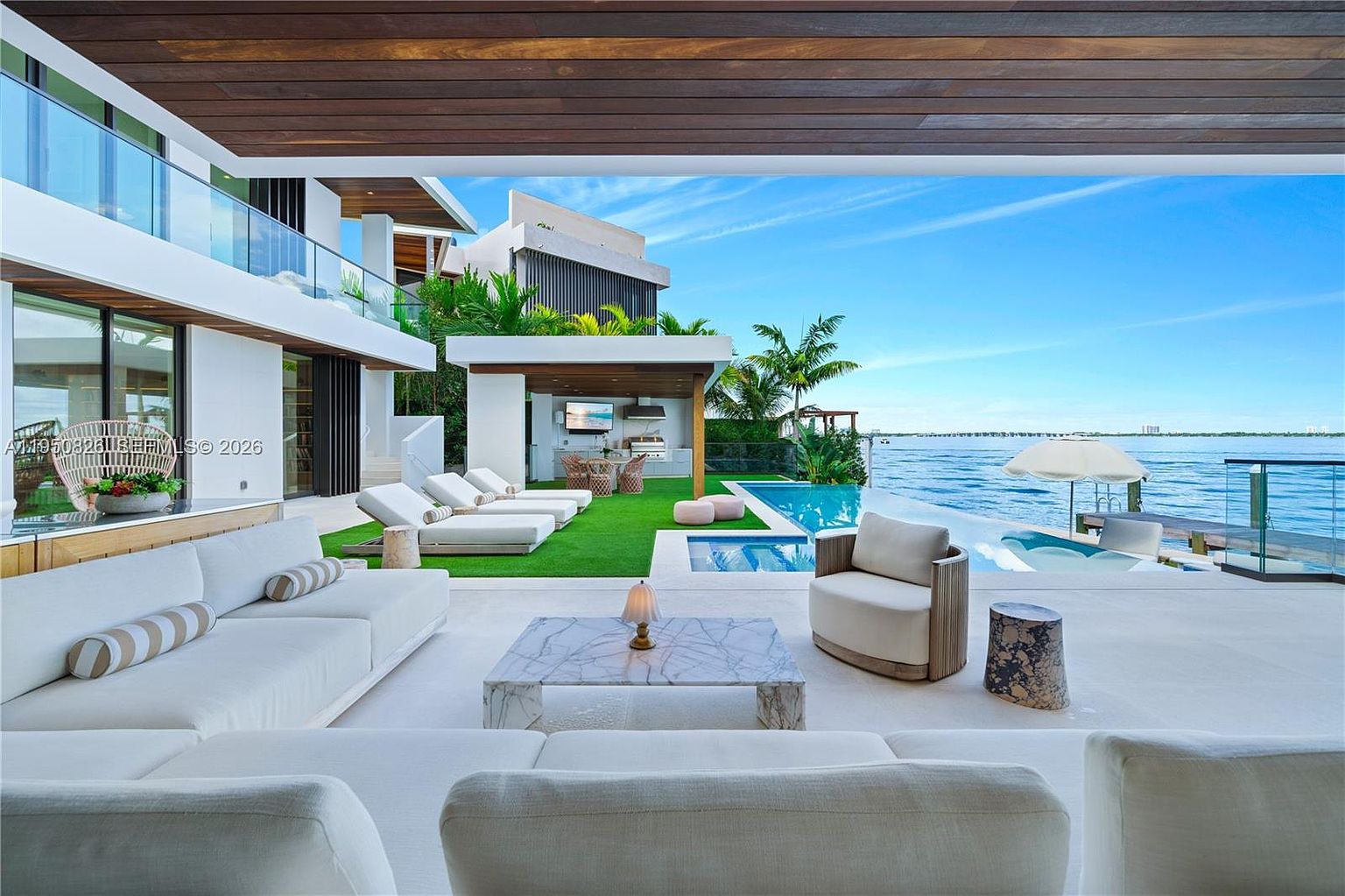 This image showcases a luxurious outdoor living space with a modern aesthetic. The patio features comfortable white sofas and a marble coffee table, leading to a pool and waterfront view. A covered outdoor kitchen and dining area are visible in the background, enhancing the property's appeal for entertaining and relaxation.