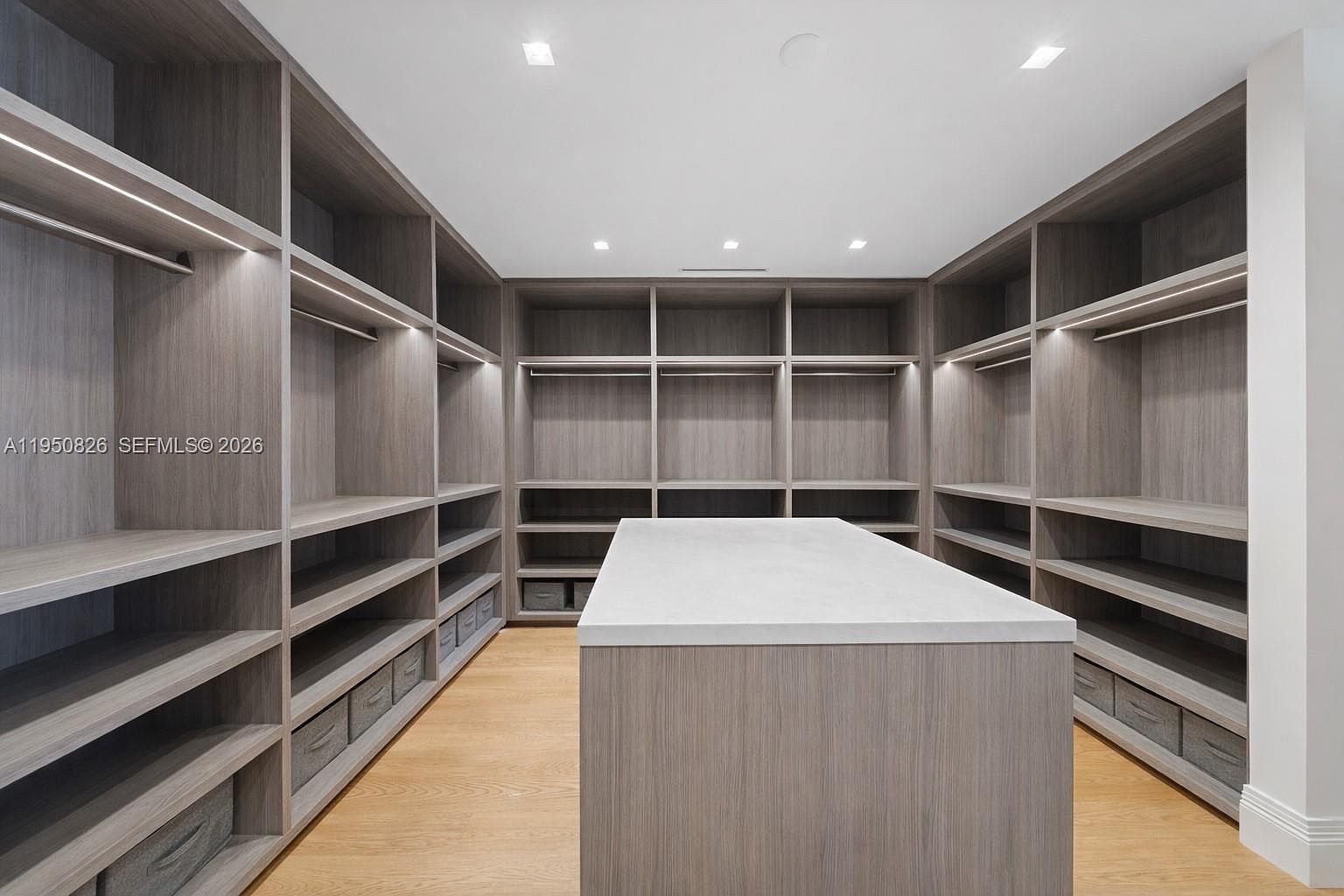 This is a spacious walk-in closet featuring custom shelving and storage solutions. The closet is well-lit with recessed lighting and under-shelf lighting, highlighting the wood-grain finish of the cabinetry. A central island provides additional surface space, enhancing the functionality and luxury of the space.