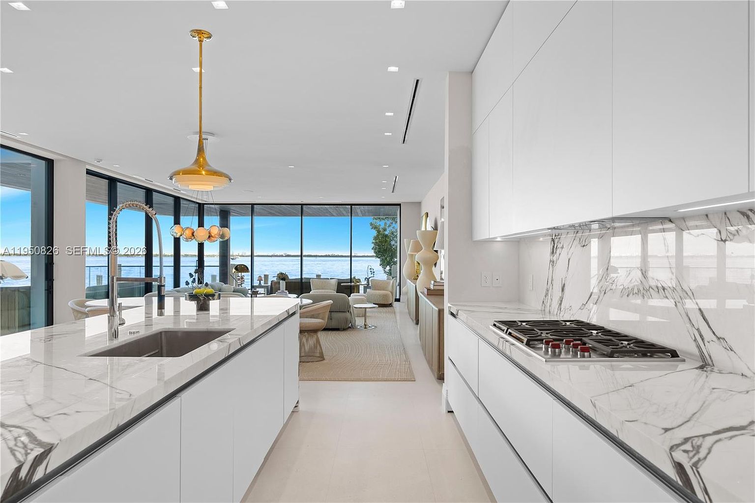 This is a bright, modern kitchen featuring sleek white cabinetry and marble countertops. A large island with a sink and faucet anchors the space, while a cooktop is integrated into the countertop along the wall. Expansive windows offer stunning water views, creating a luxurious and open atmosphere.
