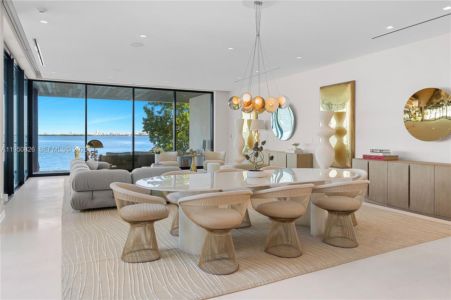 This is a bright and airy dining room featuring a large oval table with six woven chairs. A modern chandelier hangs above the table, and the room is decorated with contemporary art and vases. Large windows offer a stunning view of the water, creating a luxurious and inviting atmosphere.