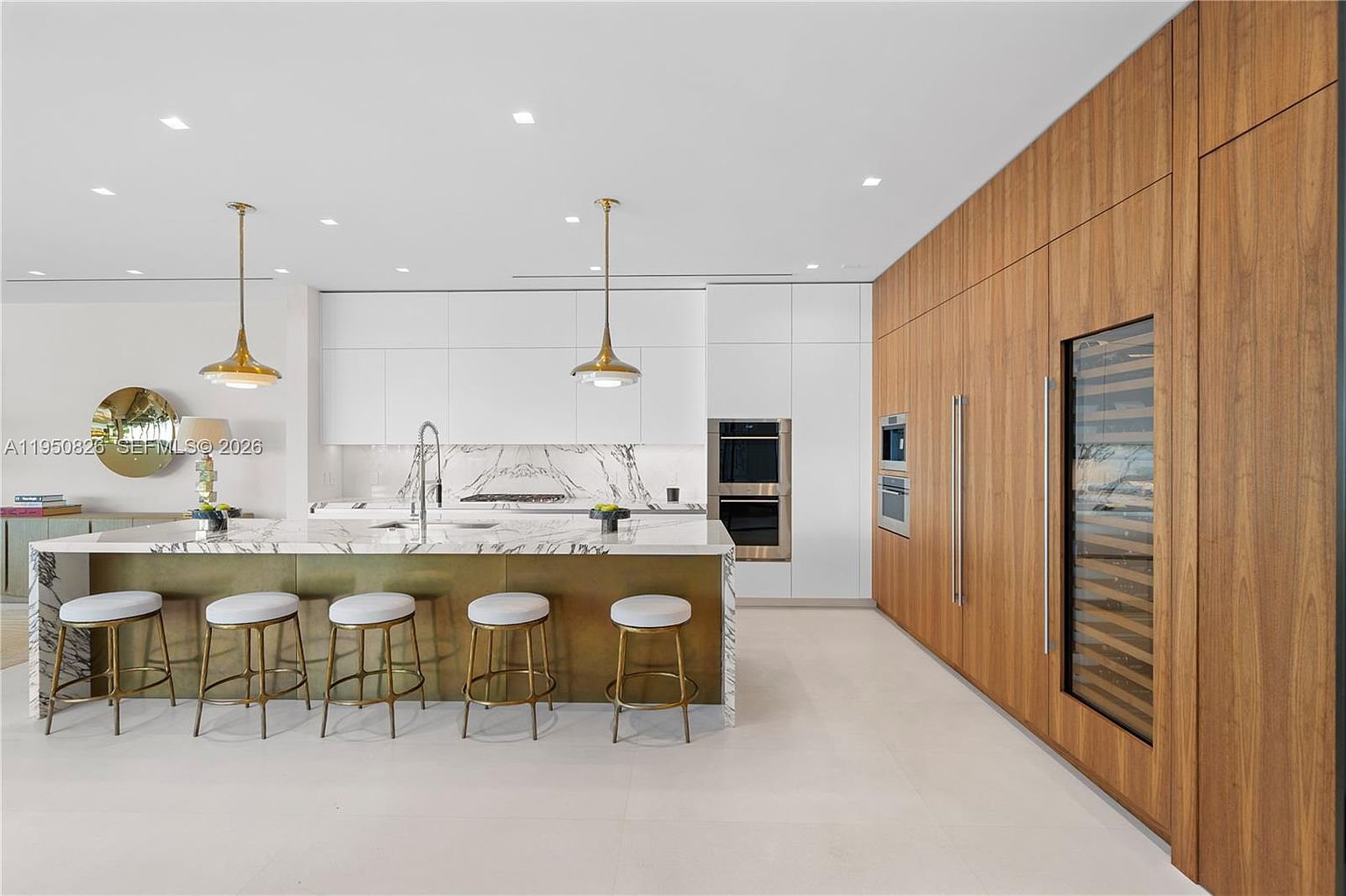This is a modern kitchen featuring a large island with seating, white cabinetry, and wood paneled walls with built-in appliances. The island has a marble countertop and gold accents, complemented by pendant lighting. The overall impression is sleek and luxurious.