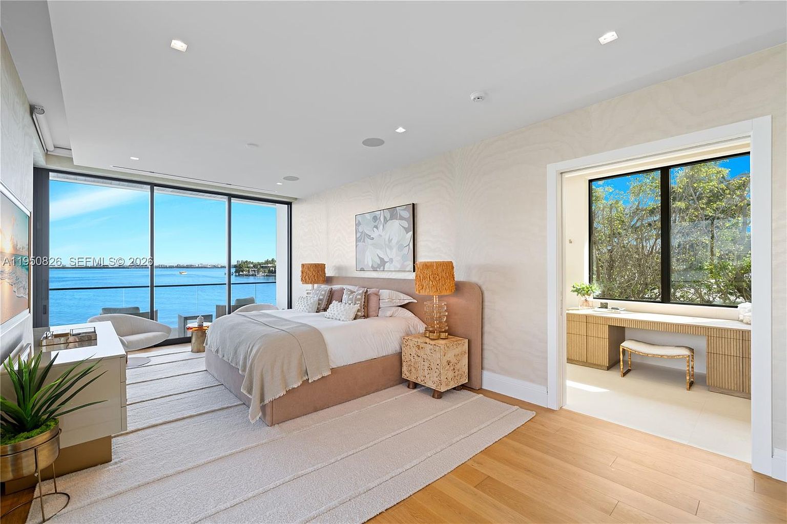 This is a luxurious primary bedroom featuring a large bed with a padded headboard, stylish nightstands with unique lamps, and a serene view of the water through expansive windows. The room is decorated in neutral tones with a textured rug and artwork, creating a calming and sophisticated atmosphere. A separate vanity area is visible through a doorway.