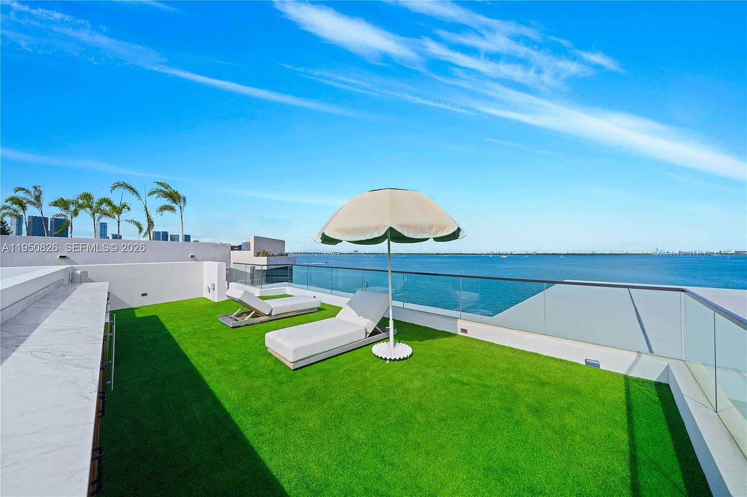 This image showcases a luxurious rooftop patio or balcony with artificial grass, two modern lounge chairs, and a large umbrella for shade. A glass railing provides unobstructed views of the ocean and cityscape in the distance. The space is designed for relaxation and entertainment, offering a private outdoor retreat.