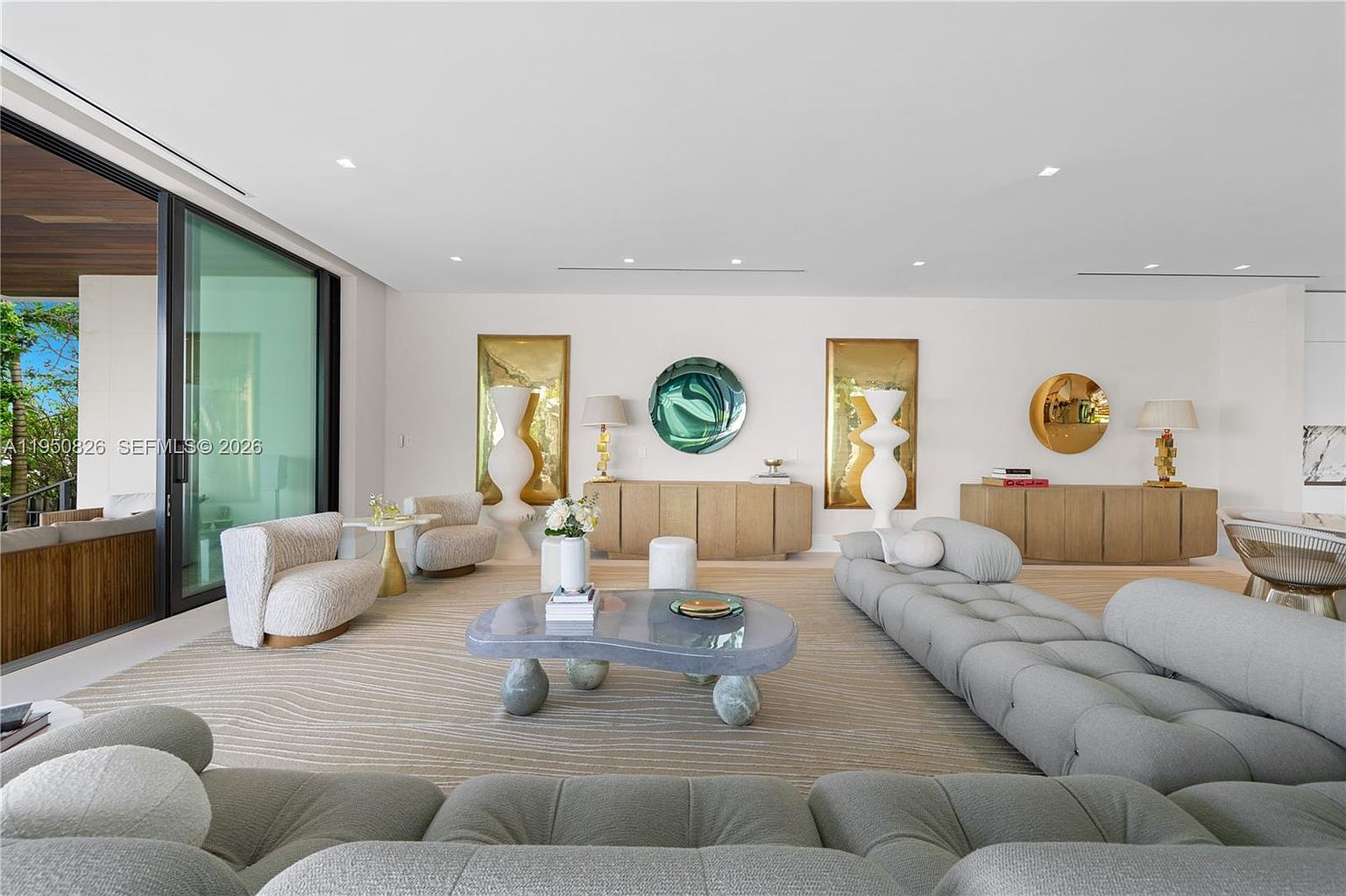 This is a spacious and modern living room featuring a neutral color palette with light gray sofas and a unique stone coffee table. The room is well-lit with natural light from a large sliding glass door and recessed lighting, complemented by stylish decor such as abstract art pieces and wooden cabinets. The overall impression is one of luxury and contemporary design.