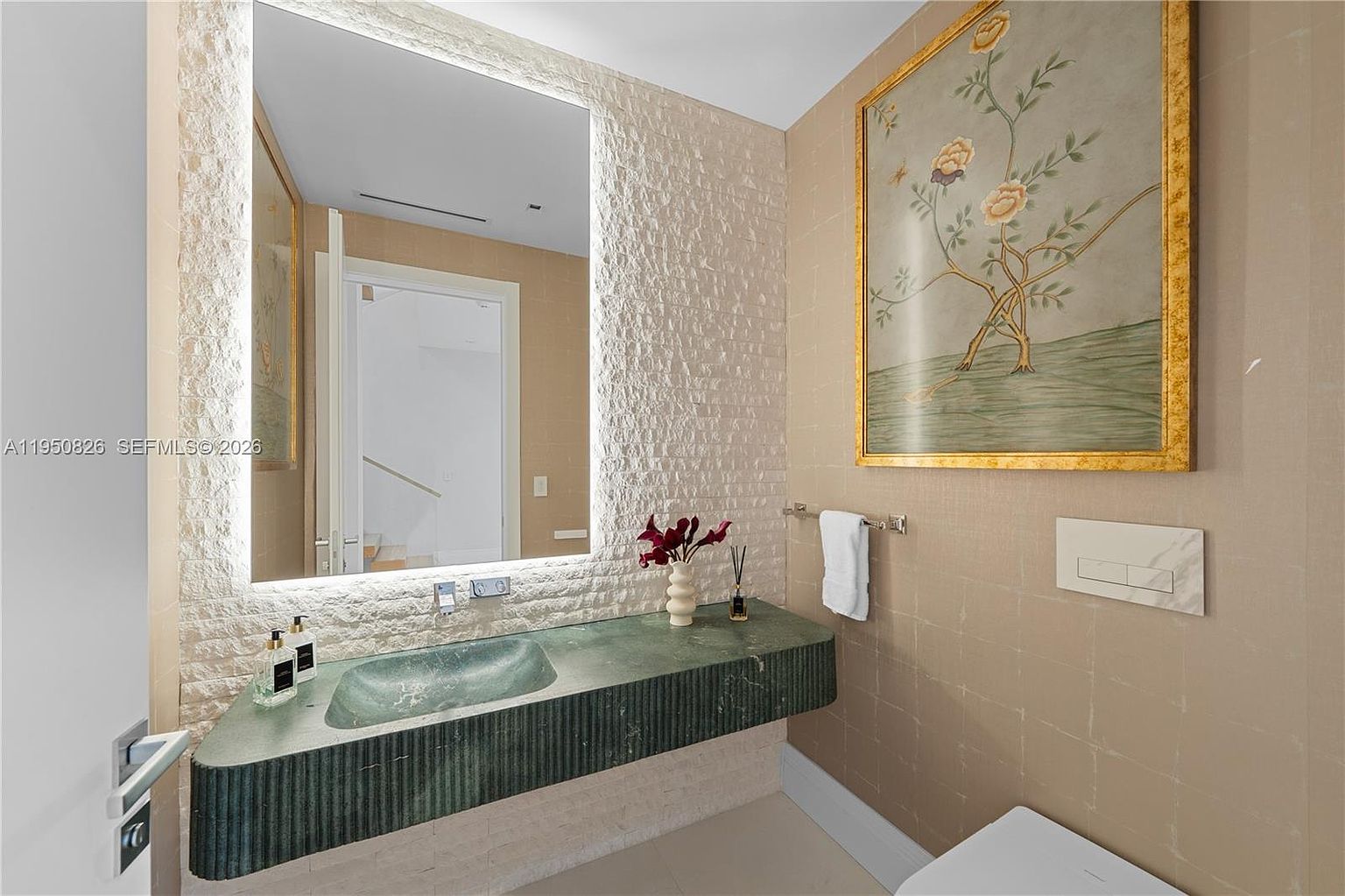 This is a well-appointed guest bathroom featuring a unique green stone sink and countertop, complemented by a large, illuminated mirror. The walls are adorned with textured tiles and a framed floral painting, adding a touch of elegance. A modern toilet and towel rack complete the space, creating a sophisticated and comfortable atmosphere.