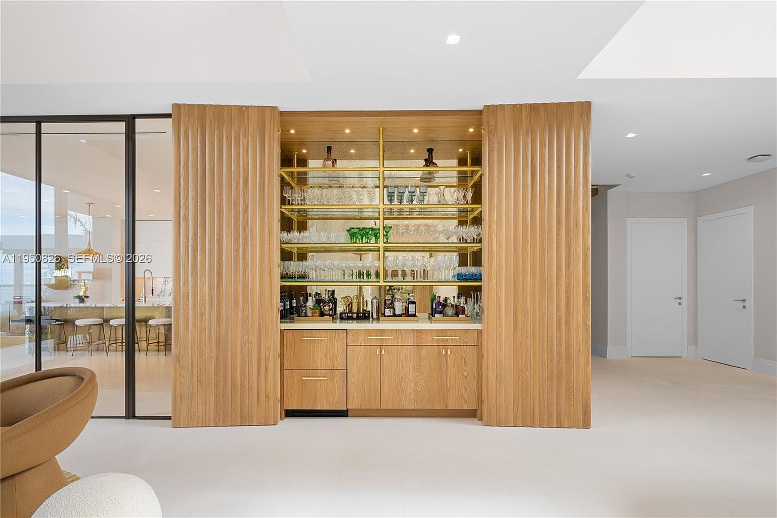 This interior shot showcases a stylish living room featuring a custom-built bar area with wooden sliding doors, gold shelving, and ample storage for glassware and beverages. The room is bright and airy, with a modern aesthetic, complemented by neutral tones and clean lines. The open layout connects to other living spaces, enhancing the sense of spaciousness.