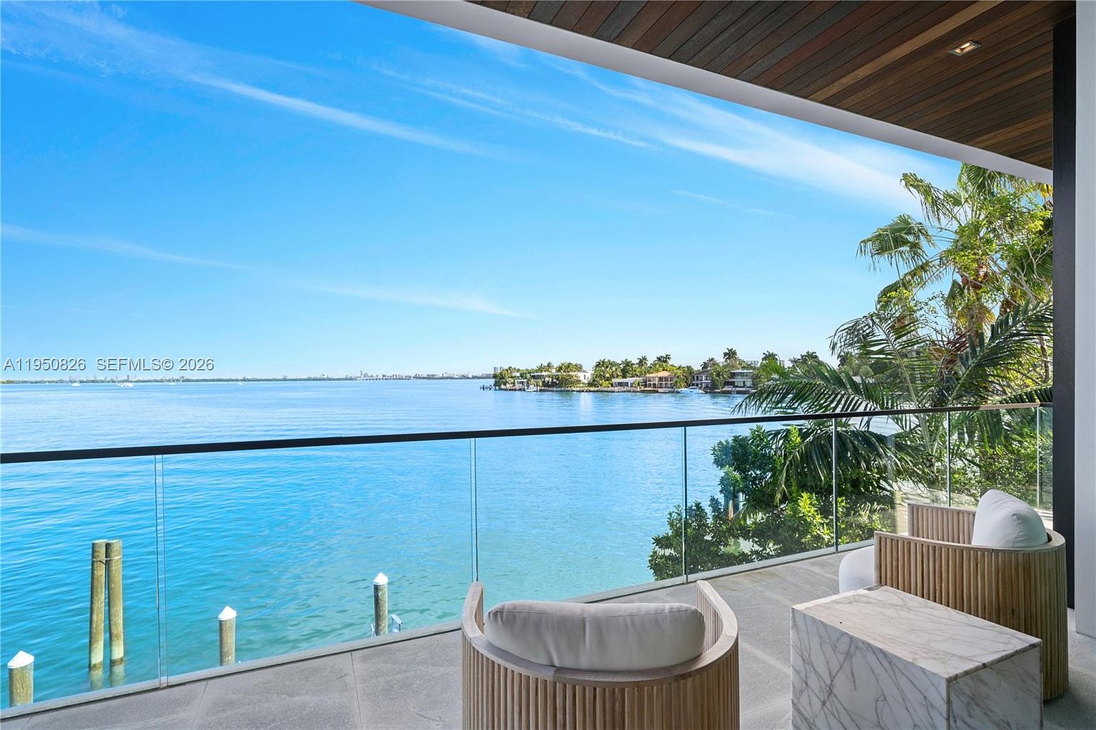 This image showcases a luxurious balcony with stunning waterfront views. The balcony features modern outdoor furniture, including stylish chairs and a marble side table, set against a backdrop of clear glass railings that offer unobstructed views of the ocean and distant cityscape. The space is designed for relaxation and entertainment, blending seamlessly with the natural beauty of the surrounding environment.
