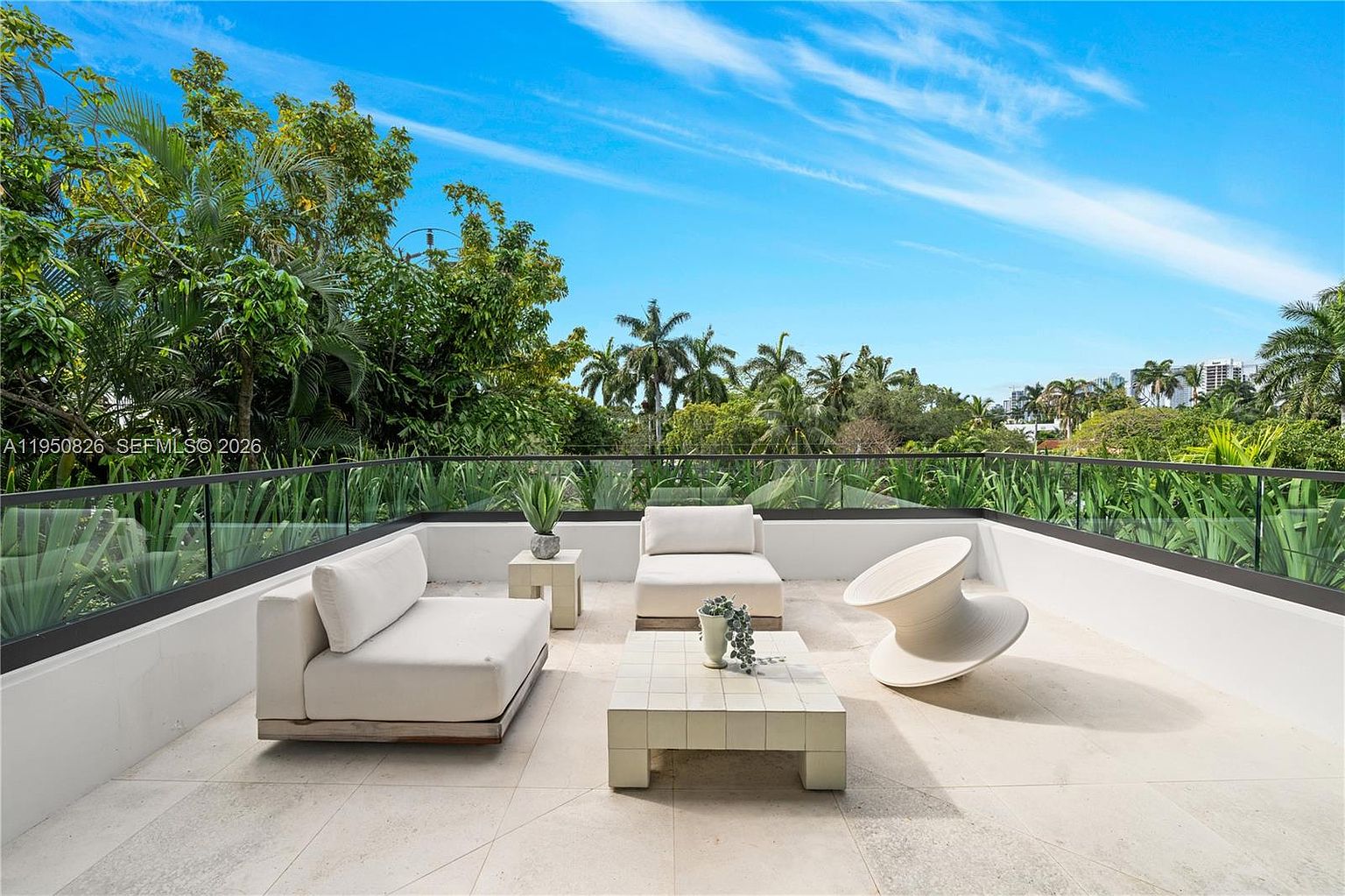 This image showcases a stylish outdoor patio or balcony area, featuring modern white furniture including a sofa, lounge chair, coffee table, and a unique sculptural piece. The space is surrounded by a glass railing, offering views of lush greenery and palm trees against a bright blue sky. The overall impression is one of luxury and relaxation, perfect for outdoor entertaining or enjoying the natural surroundings.