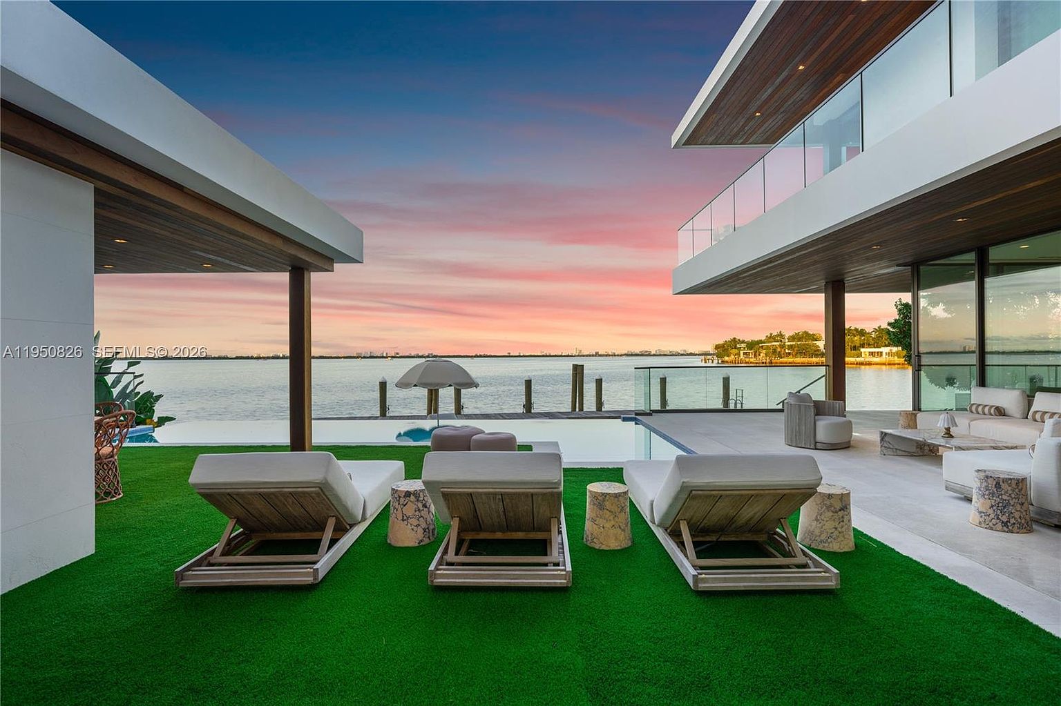 This exterior shot showcases a luxurious pool area at sunset, featuring modern architecture with clean lines and wood accents. Three lounge chairs are arranged on the artificial turf, overlooking the infinity pool and the bay. The scene exudes tranquility and upscale living, perfect for relaxation and entertainment.