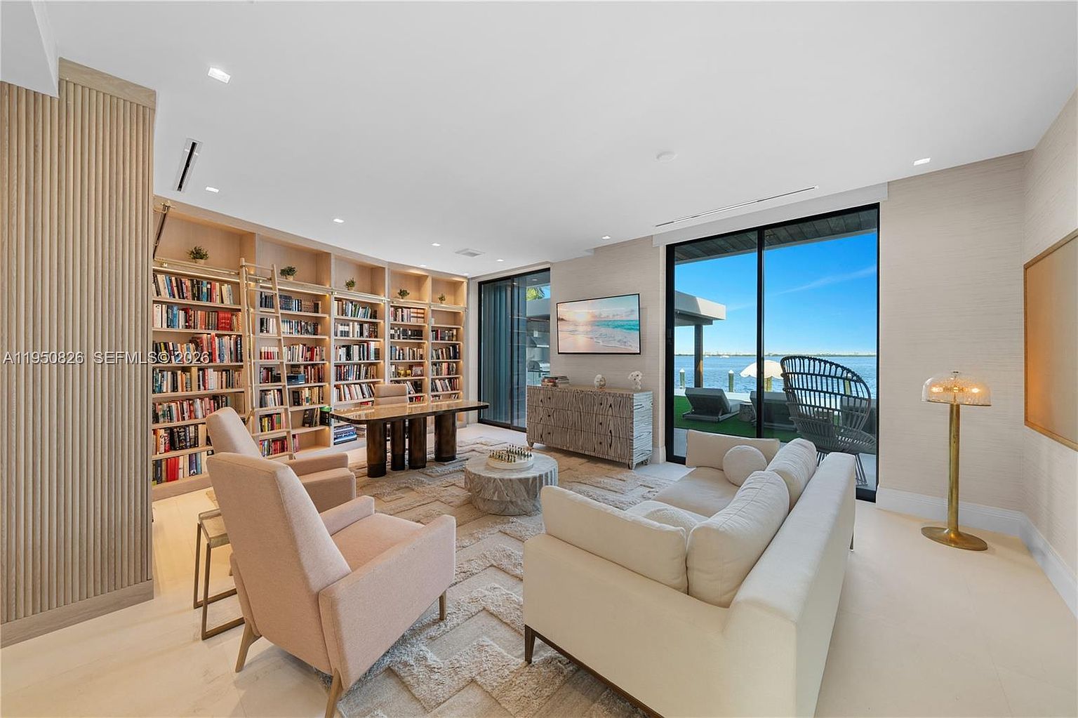 This is a spacious and luxurious living room featuring a large bookshelf filled with books, a modern desk, and comfortable seating including armchairs and a sofa. The room has large windows offering a view of the waterfront, and the decor is sophisticated with a neutral color palette and textured walls. The overall impression is one of elegance and comfort.