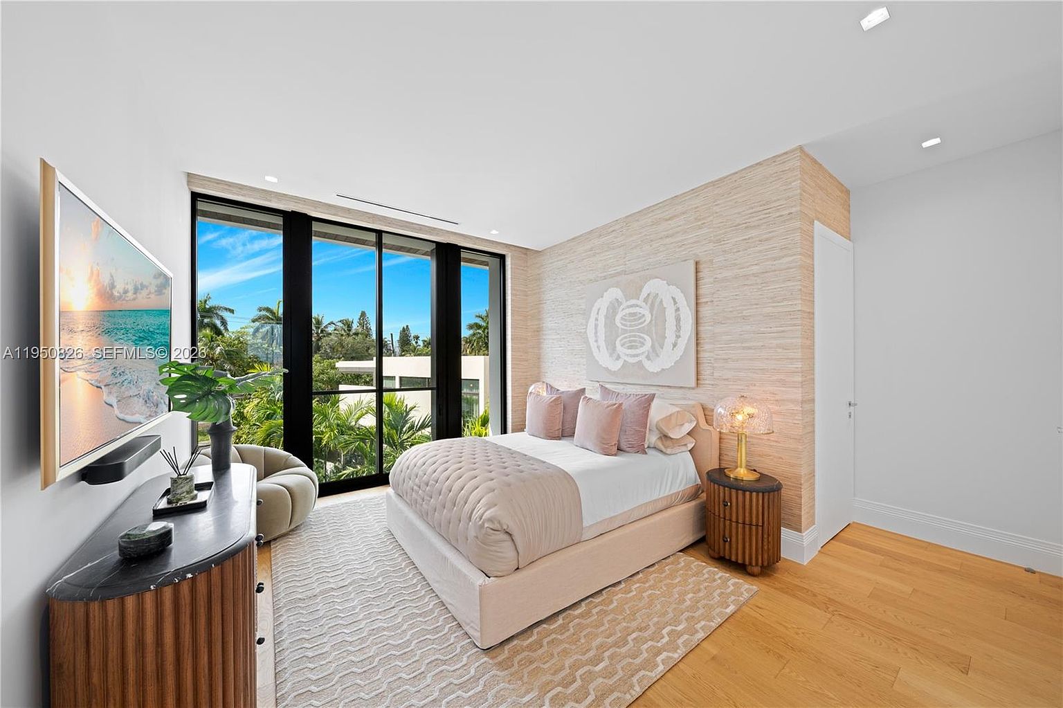This is a bright and airy primary bedroom featuring a large window with a view of lush greenery. The room is decorated in neutral tones with a textured accent wall and a light-colored bed with pink pillows. A modern dresser with a TV and a round chair are placed near the window, and a wooden nightstand with a lamp sits beside the bed, creating a serene and inviting atmosphere.