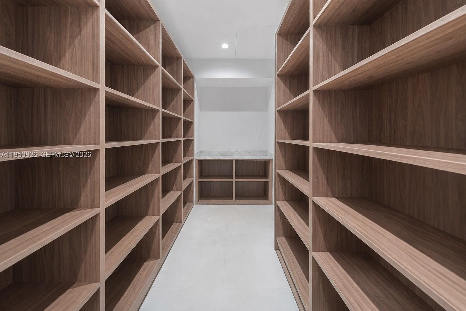 This is a well-organized walk-in closet featuring custom wooden shelving on both sides of a central aisle. The shelves are empty, suggesting ample storage space. At the end of the closet, there is a built-in unit with a marble countertop, adding a touch of luxury to the space.