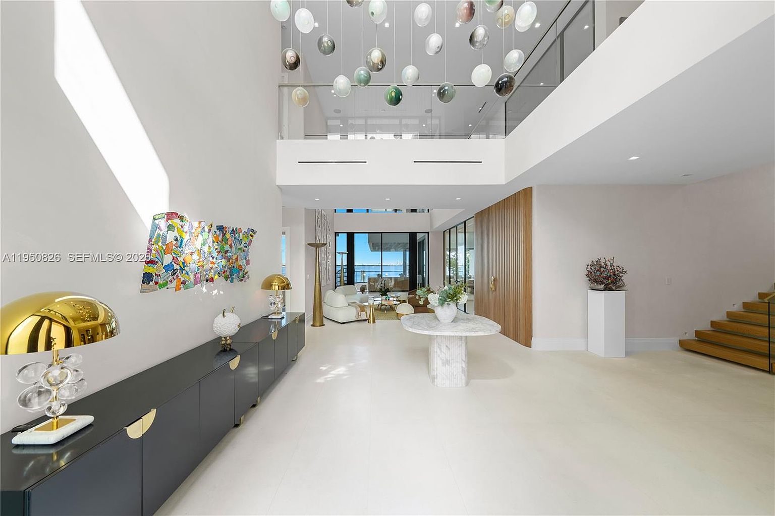 This is a grand foyer featuring high ceilings and an open floor plan. The space is decorated with modern art and lighting fixtures, creating a luxurious and inviting atmosphere. A staircase with glass railings is visible on the right, leading to the upper level.
