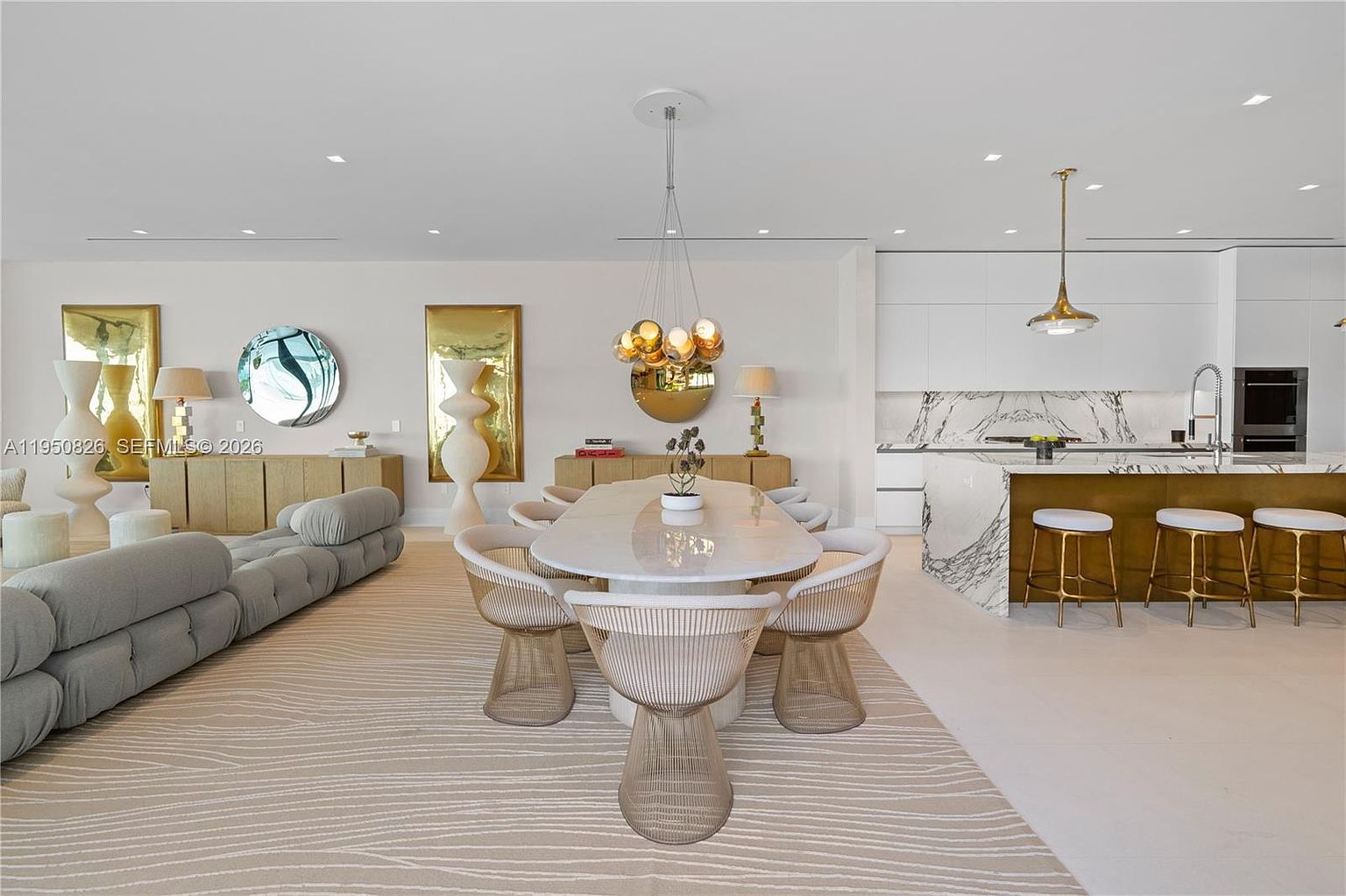 This is an open-concept living and dining area with a modern aesthetic. The living room features a large, comfortable sofa and a patterned rug, while the dining area boasts a sleek, oval table surrounded by stylish chairs and a unique chandelier. The kitchen is visible in the background, showcasing white cabinetry and marble countertops, creating a cohesive and luxurious living space.