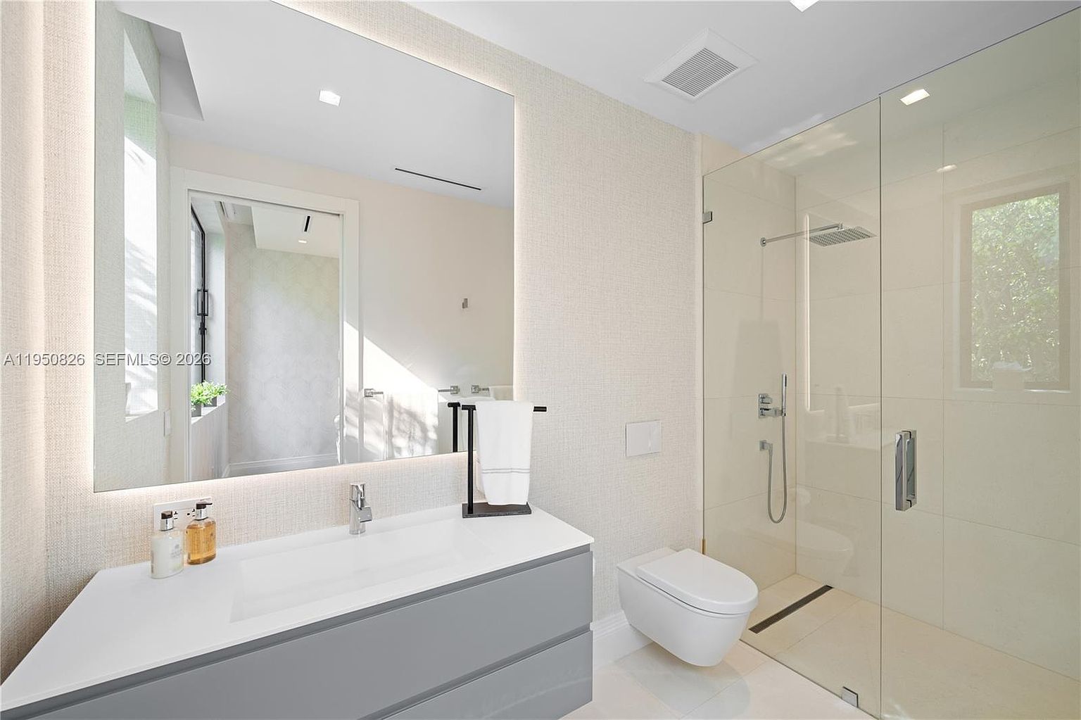 This is a modern primary bathroom featuring a floating vanity with a gray base and white countertop, a large mirror with integrated lighting, and a glass-enclosed shower with a rainfall showerhead. A wall-mounted toilet adds to the sleek design, and the neutral color palette creates a spa-like atmosphere. The perspective is from the entrance of the bathroom, showcasing the vanity and shower area.