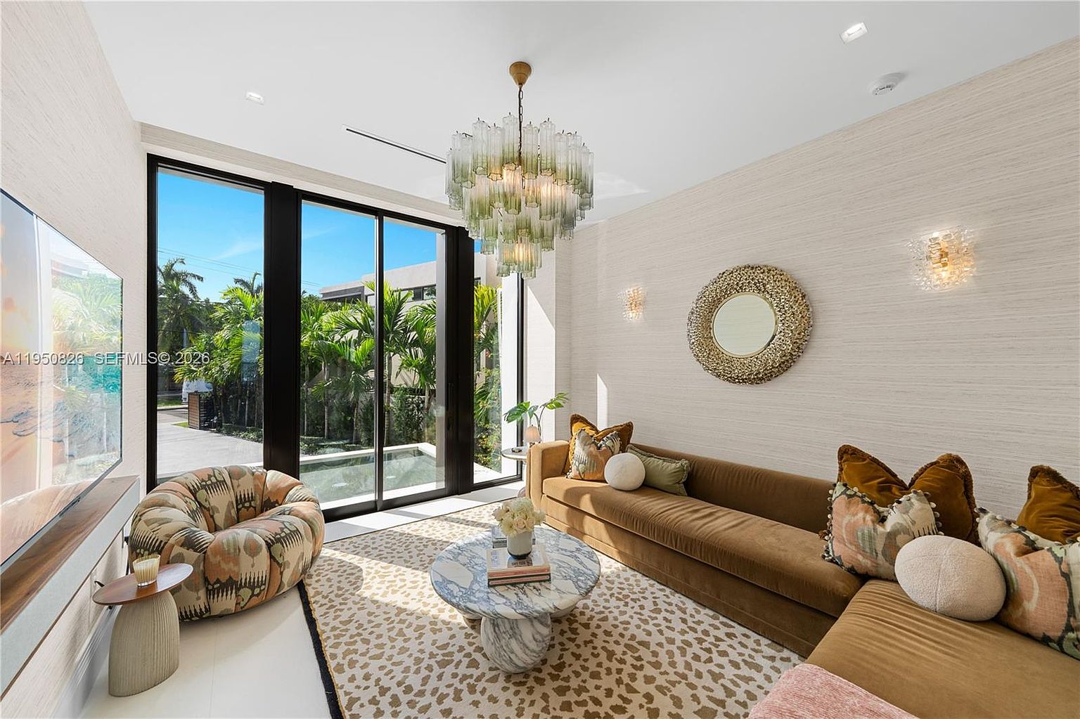 This is an interior shot of a stylish living room featuring a brown sectional sofa adorned with decorative pillows, a round marble coffee table, and a unique patterned rug. A modern chandelier hangs from the ceiling, and large windows offer a view of lush greenery outside. The room exudes a sophisticated and comfortable ambiance.