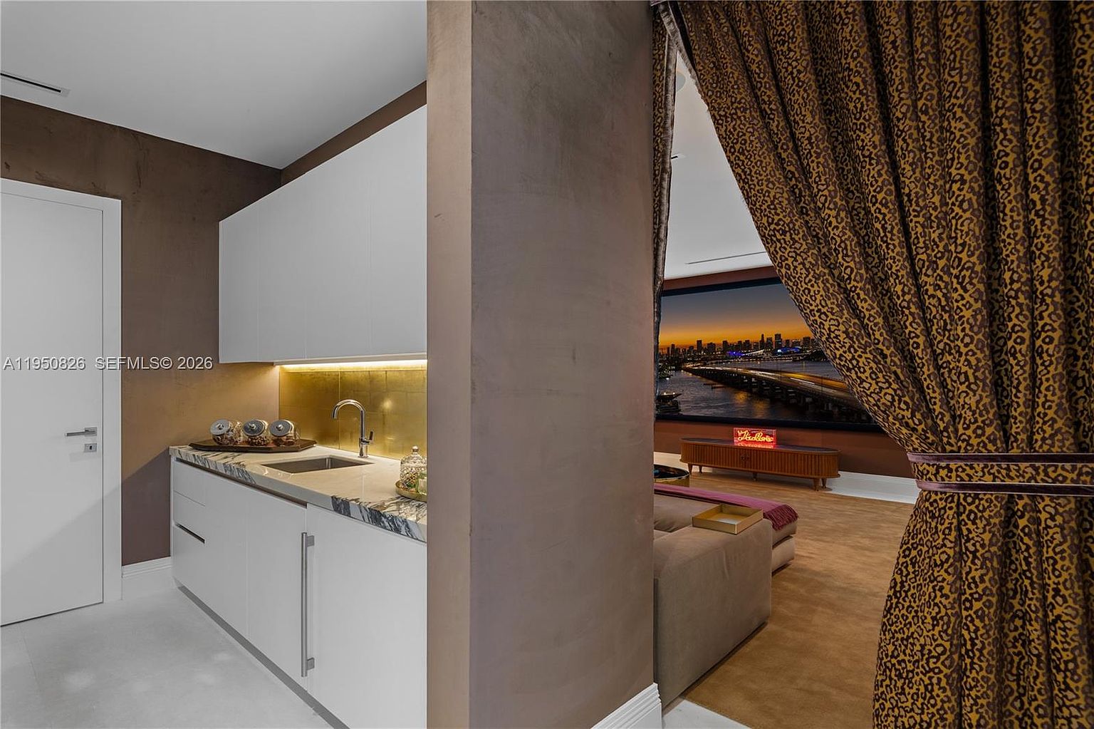 This interior shot showcases a modern kitchen area with sleek white cabinetry and a marble countertop. A sink and faucet are visible, along with decorative items on the counter. Through an opening framed by a decorative curtain, a glimpse of a living area with a cityscape view can be seen, adding depth and luxury to the space.