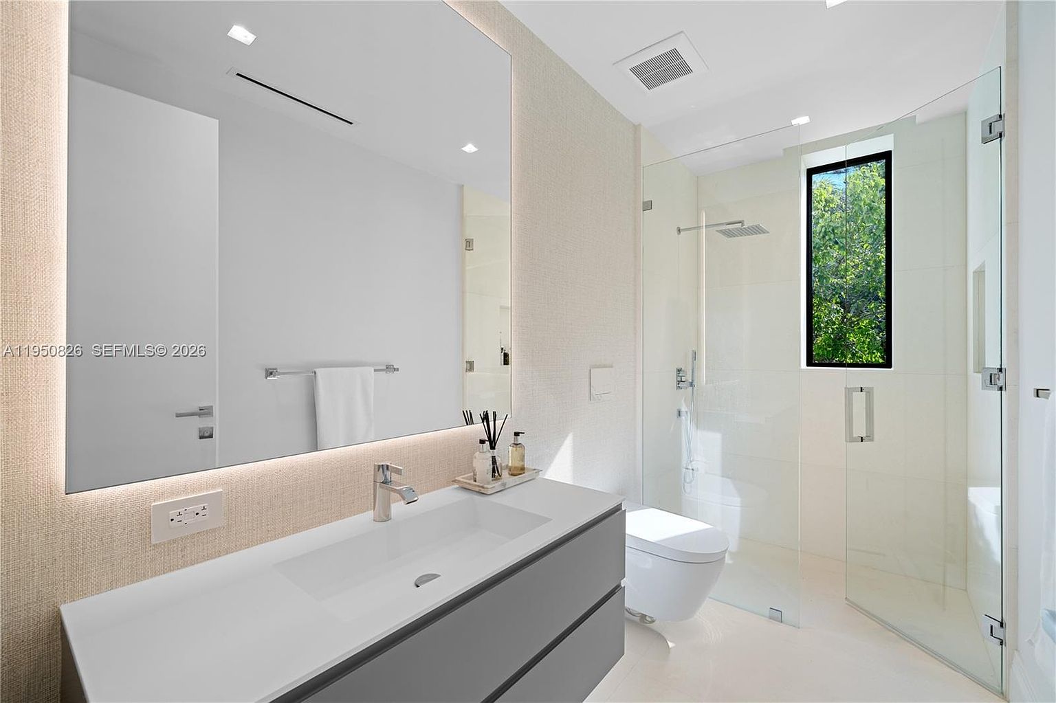 This is a modern primary bathroom featuring a large rectangular mirror with integrated lighting above a sleek, gray vanity with a white countertop and integrated sink. The bathroom includes a glass-enclosed shower with a window offering natural light and a view of greenery. The overall design is minimalist and luxurious, with clean lines and a neutral color palette.