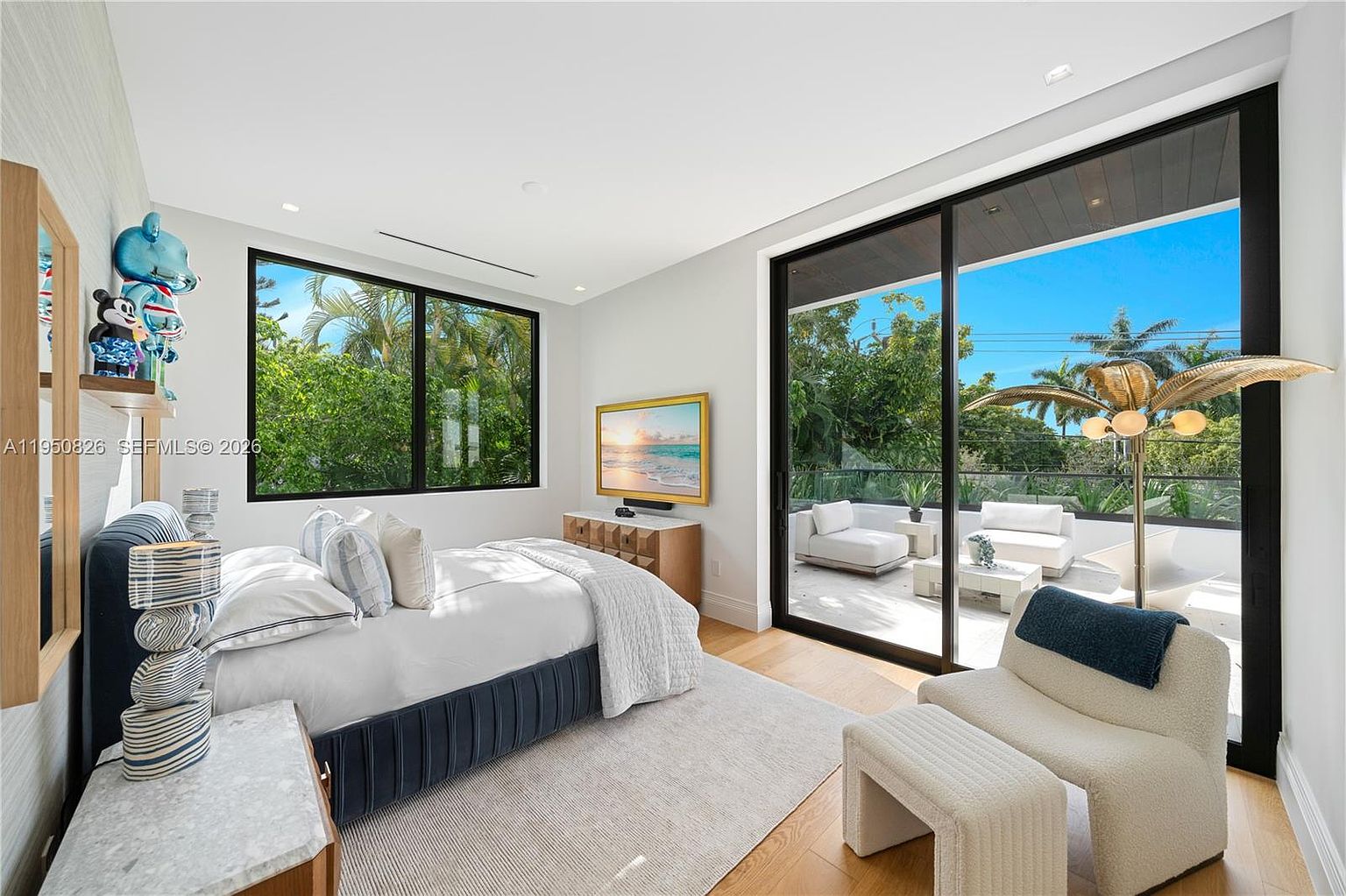 This is a bright and airy primary bedroom featuring a large bed with a blue upholstered base and white linens. A large window and sliding glass doors offer views of lush greenery and a private balcony with outdoor seating. The room is decorated in a modern style with a neutral color palette and natural wood accents.