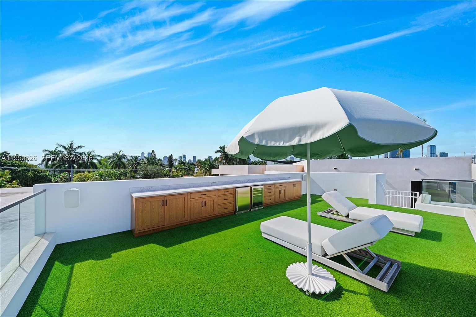This image showcases a luxurious rooftop patio featuring artificial turf, a built-in outdoor kitchen with wooden cabinetry and a stainless steel refrigerator, and two white lounge chairs under a large white umbrella. The patio is surrounded by a white wall with glass railings, offering views of the city skyline and lush greenery in the distance. The bright blue sky and modern design create an inviting and upscale outdoor living space.