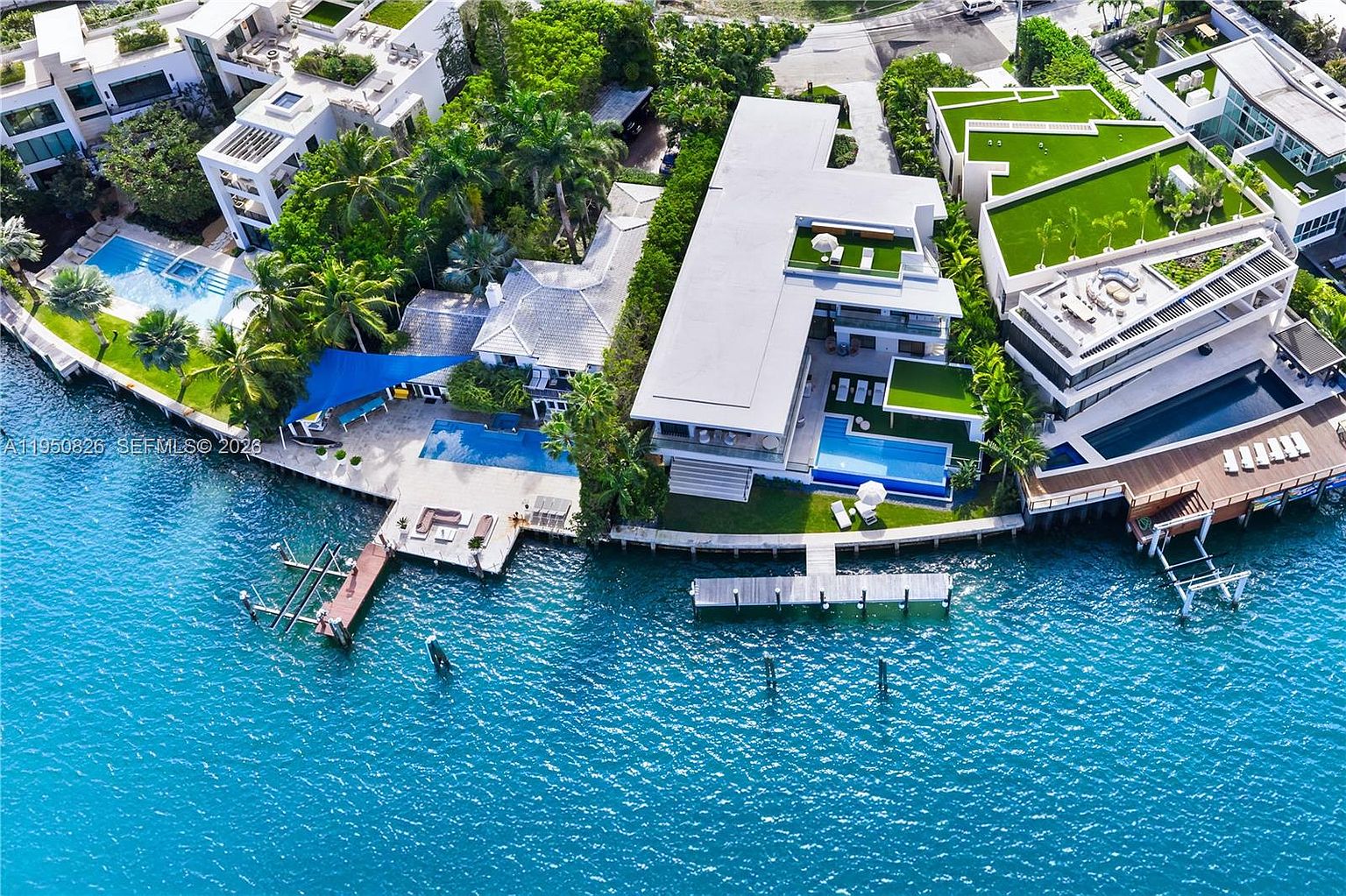 This aerial shot showcases a luxurious waterfront property featuring multiple modern homes with flat roofs, lush landscaping, and private docks. Several swimming pools and outdoor lounge areas are visible, emphasizing the property's high-end amenities and prime location. The turquoise water surrounding the estate adds to the overall impression of exclusivity and tropical paradise.