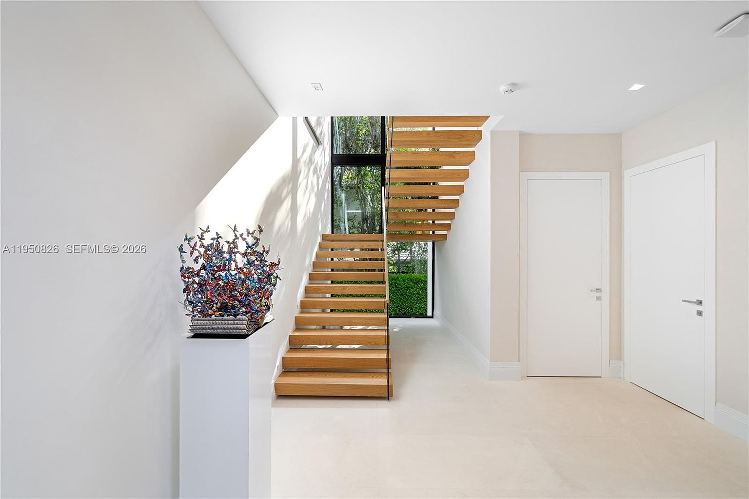 This interior shot showcases a modern hallway with floating wooden stairs leading upwards. A striking butterfly sculpture sits atop a white pedestal, adding an artistic touch. Natural light streams in through a window beside the stairs, highlighting the clean lines and minimalist design of the space, which includes two white doors.