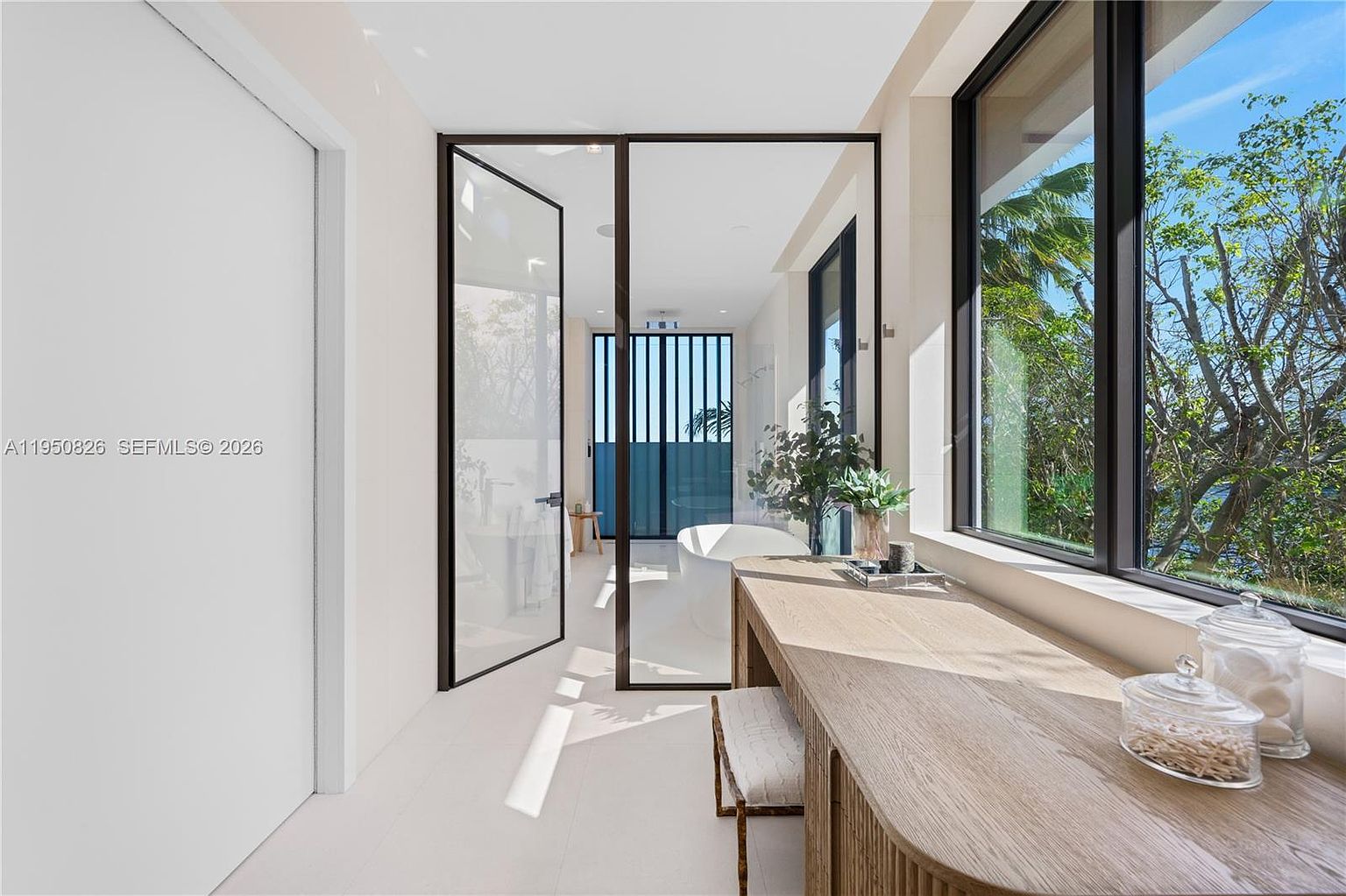 This is a bright and modern primary bathroom featuring a sleek design with a large window offering natural light and a view of the outdoors. The bathroom includes a vanity area with a wooden countertop, a stylish stool, and decorative jars. A glass-enclosed area likely contains a shower or tub, enhancing the spa-like atmosphere.