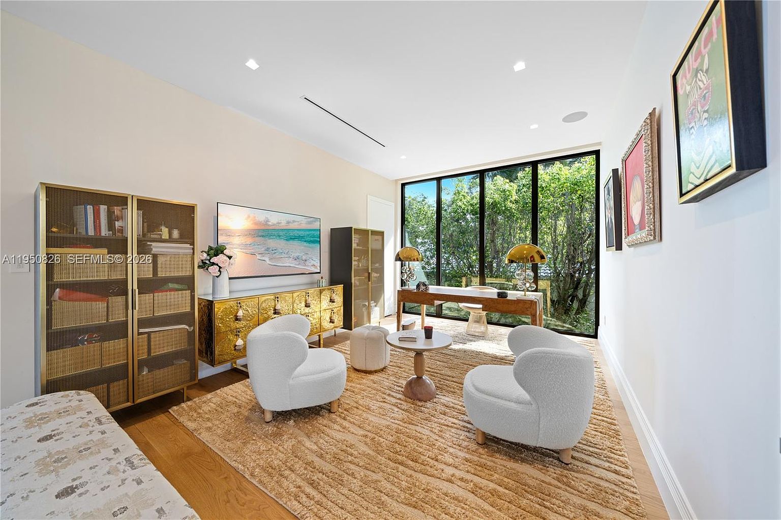 This interior shot showcases a modern office space featuring a large window with a view of lush greenery. The room is furnished with two white armchairs, a small round table, a wooden desk, and a gold-accented cabinet, creating a stylish and functional workspace. The overall aesthetic is bright, airy, and conducive to productivity.