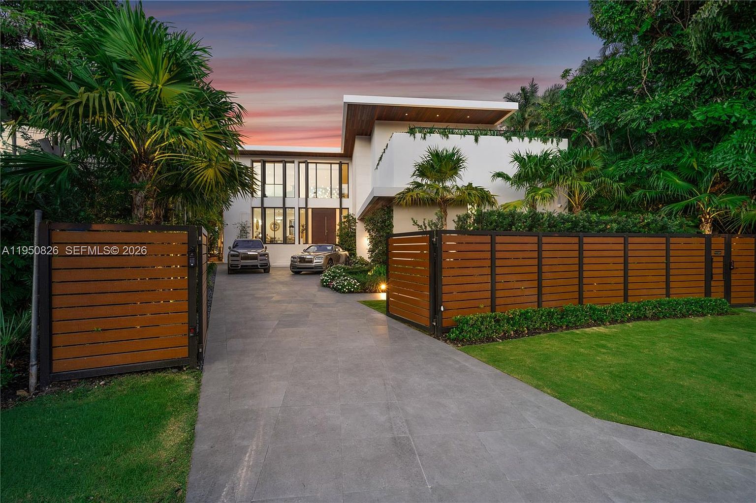 This is a front exterior view of a modern luxury home. The property features a gated entrance with a long driveway leading to the house, showcasing two luxury cars parked in front. The architecture is contemporary with clean lines, a flat roof, and large windows, complemented by lush landscaping and mature trees, creating a sense of privacy and exclusivity.